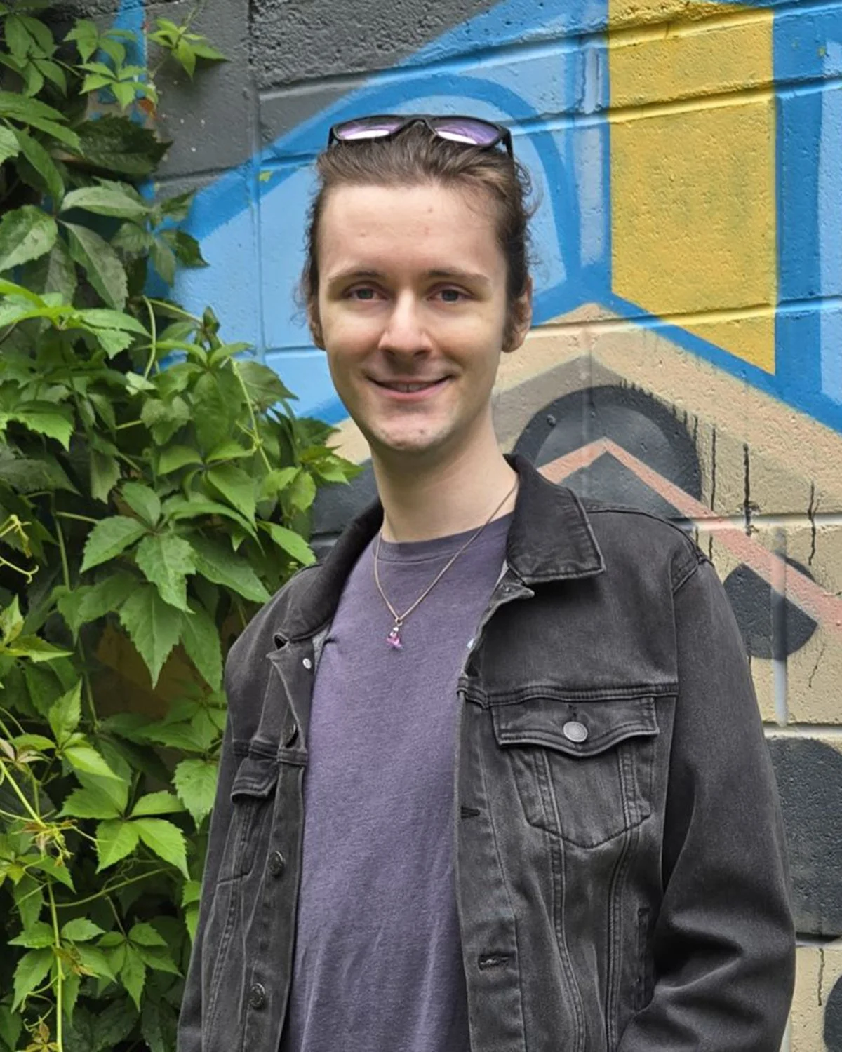 A young man with short brown hair, wearing sunglasses on his head, a gray t-shirt, and a black denim jacket, standing outdoors in front of a colorful graffiti mural and green leafy plants, smiling at the camera.