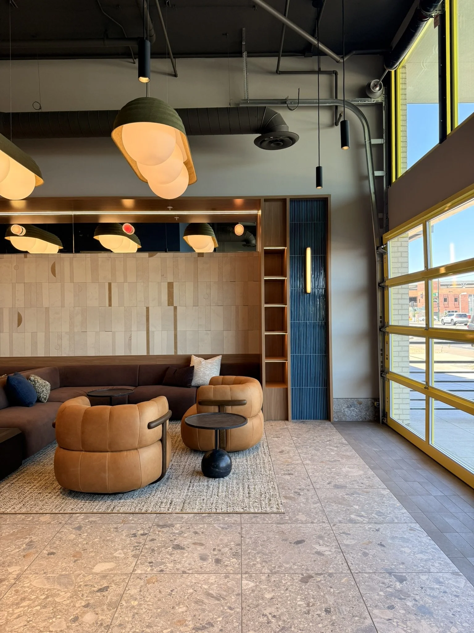 Modern cafe interior with a brown sofa, an orange leather ottoman, black side tables, artwork, wood shelving unit, and large windows with yellow frames, bright natural light, and views outside.