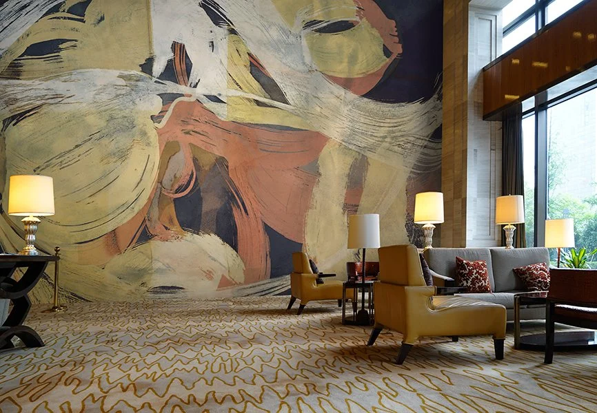 Interior of a modern hotel lobby with abstract wall art, large windows, beige and yellow furniture, and multiple lamps.