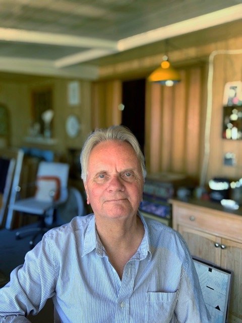 Man standing in art studio with colorful abstract painting behind him, worktable with paint cans and art supplies, and windows with natural light.