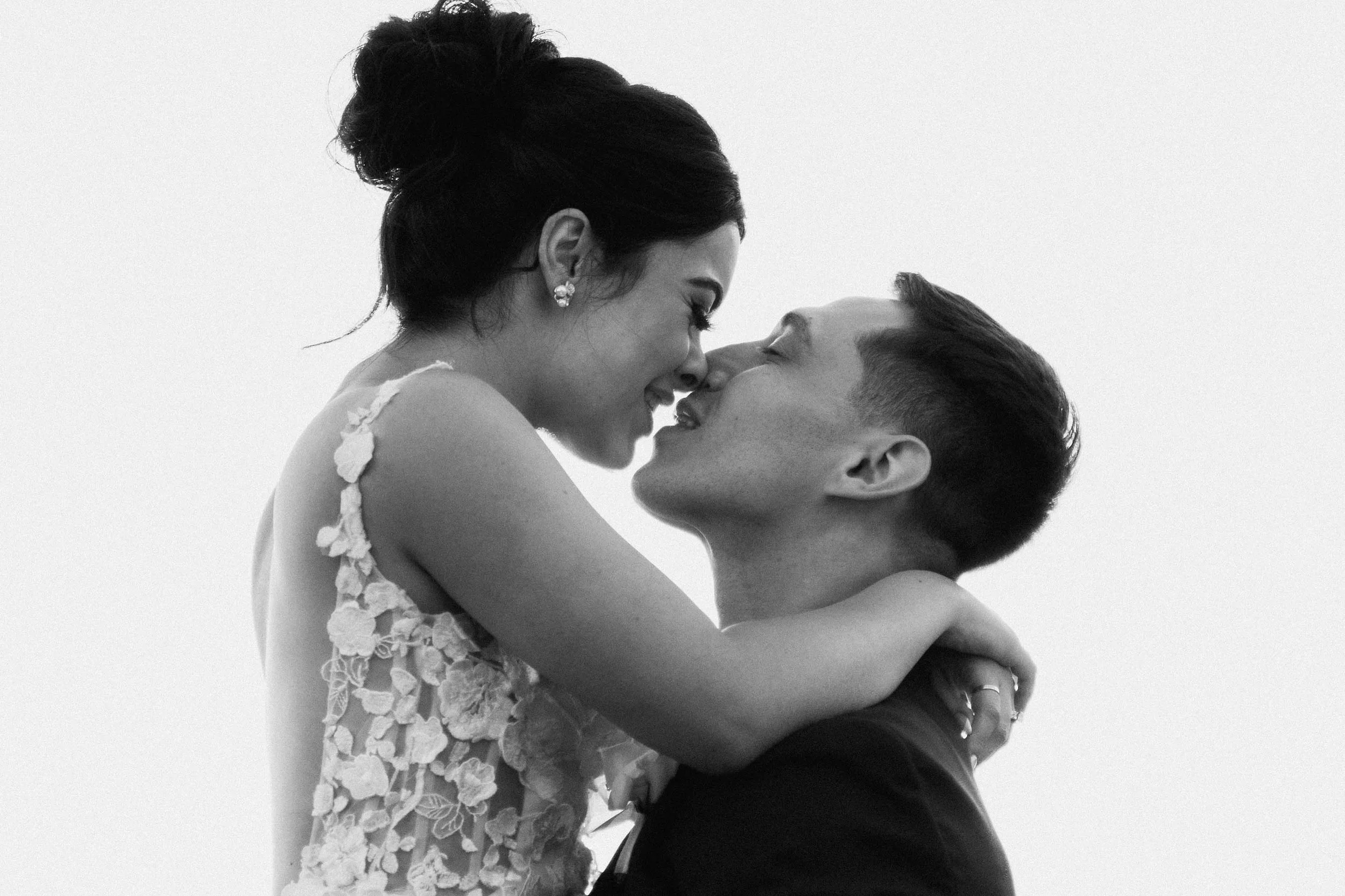 A woman wraps her arms around a man as they lean together mid-kiss, framed closely like a cinematic prenup film still.