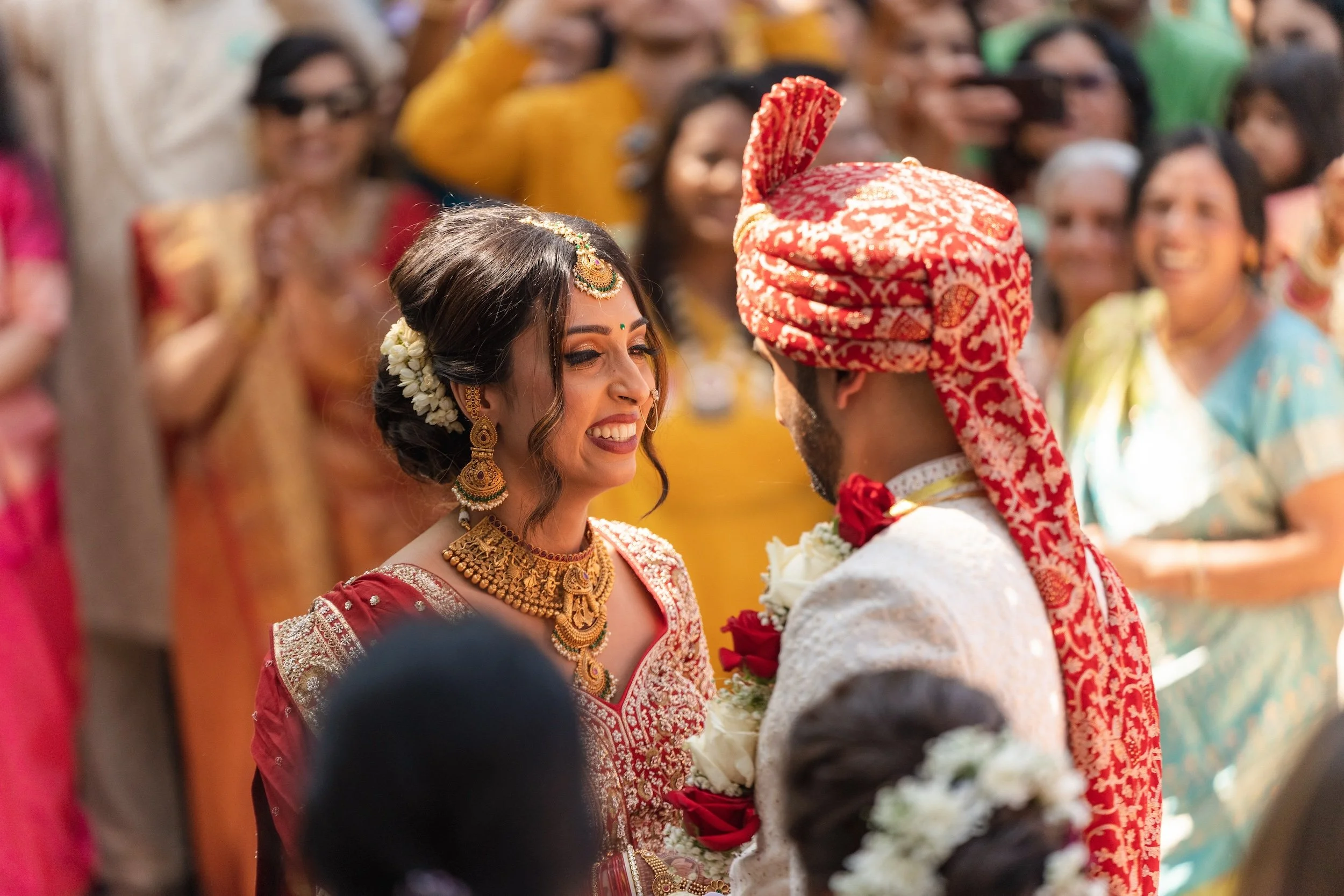 Bride and groom smiling at each other during their ceremony, framed by family and golden light — captured by Caz Isaiah, India Wedding Photographer