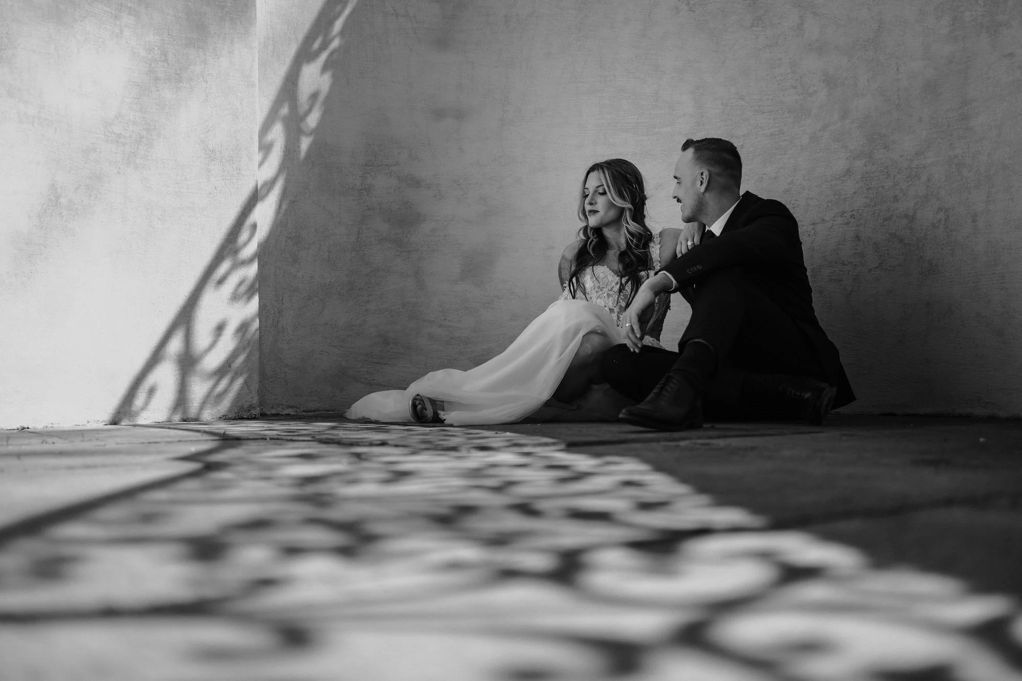 Bride and groom seated together against a textured wall, shadows falling across them in an intimate monochrome portrait by Caz Isaiah.