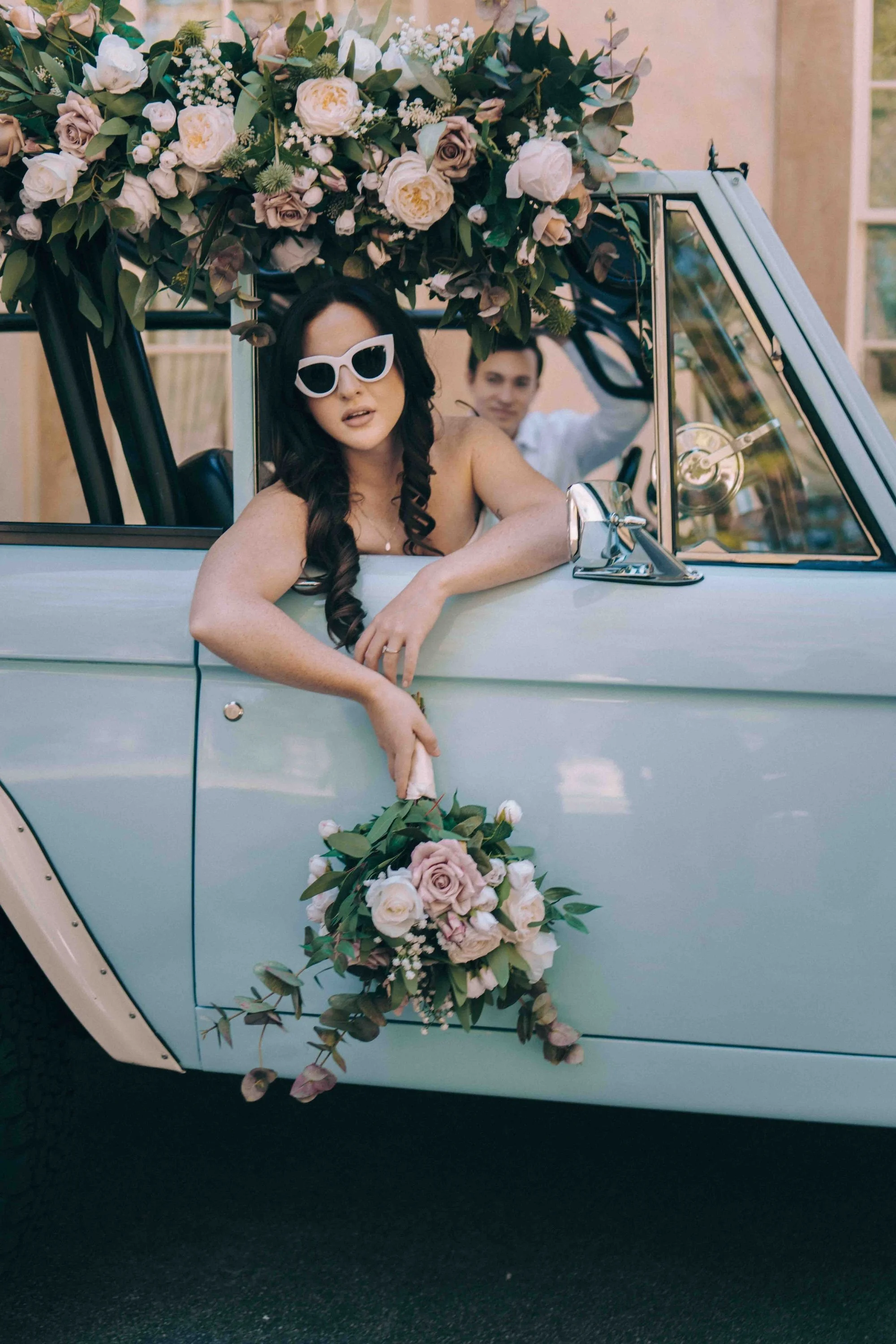 A bride leans playfully out of a vintage car window, bouquet in hand, sunglasses adding a cinematic flair to the scene.
