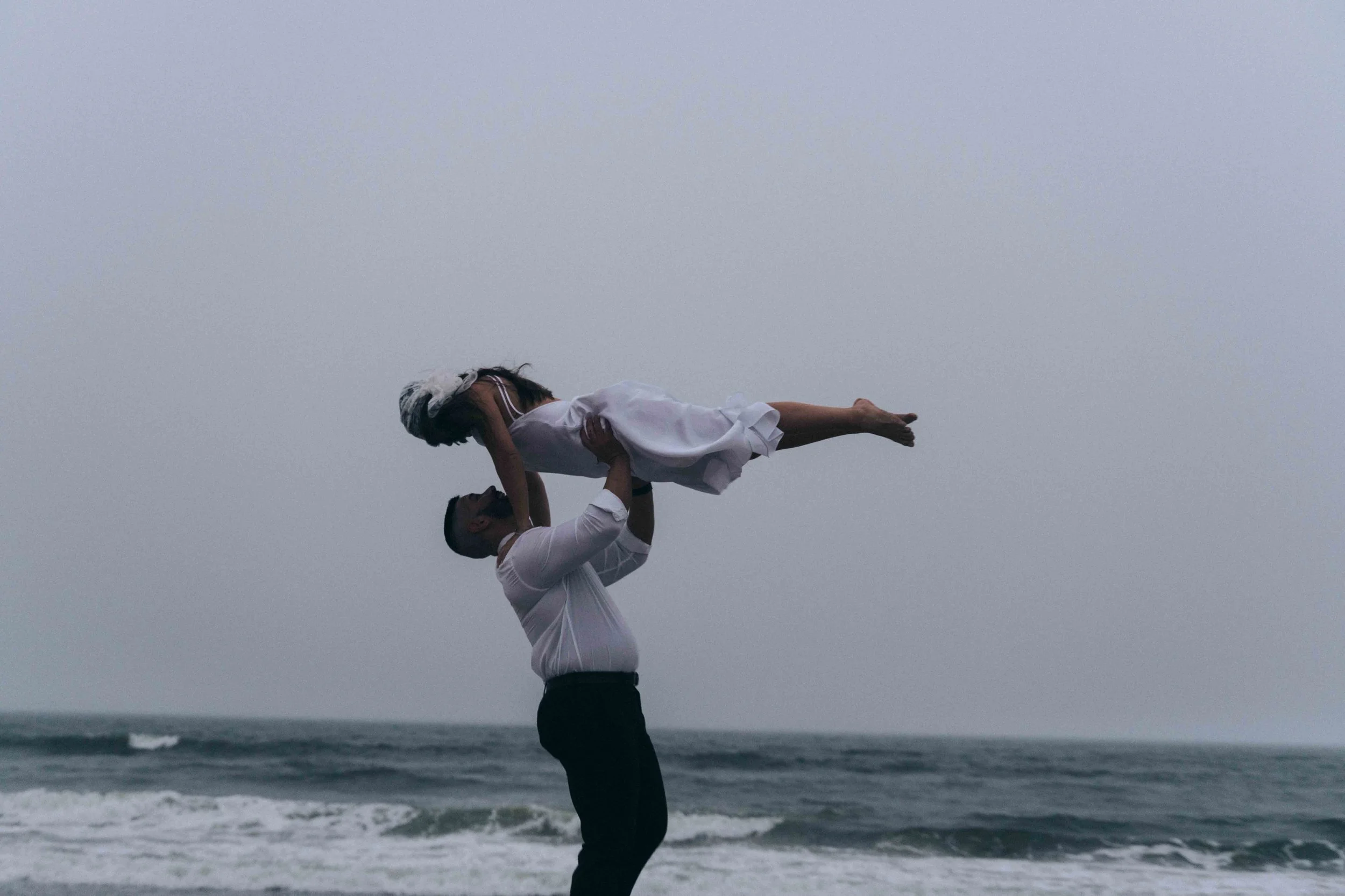 A man lifts a woman above him at the ocean’s edge, her dress extended midair like a cinematic prenup film frame.