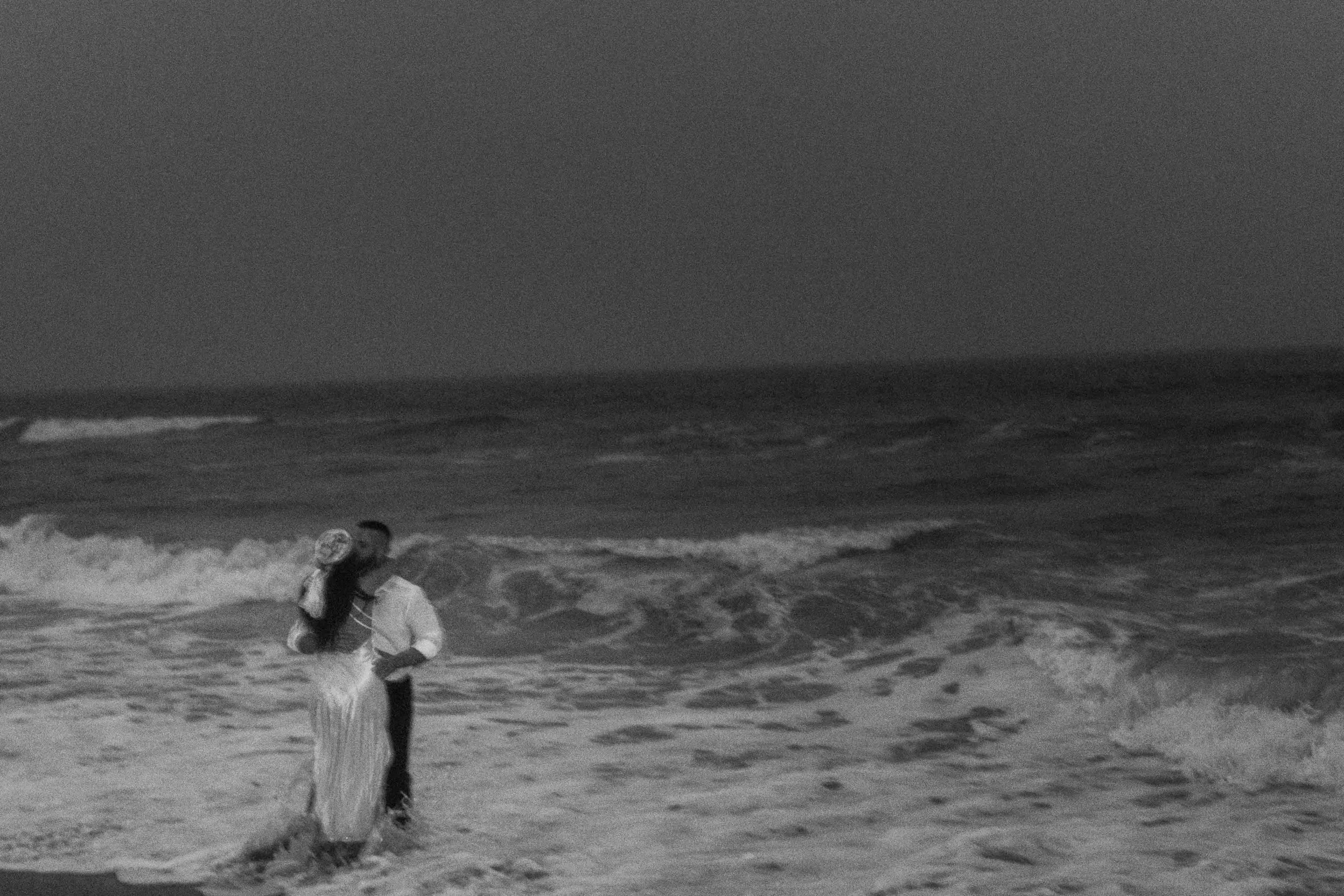 A couple walks side by side along dark sand near the waterline, their figures moving slowly as the shoreline recedes behind them.
