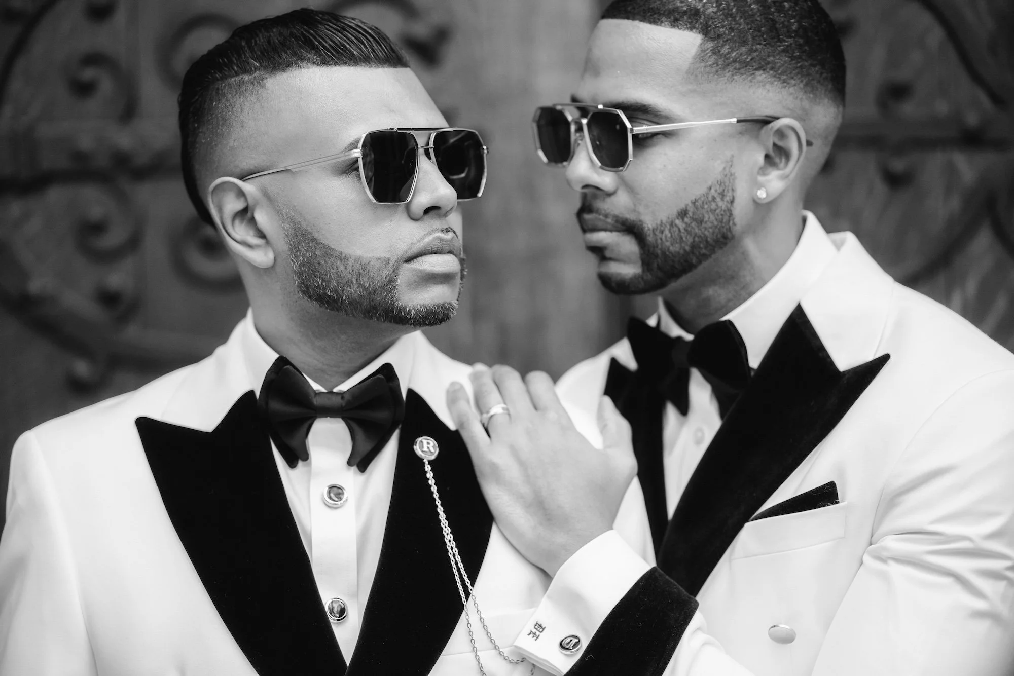 Two grooms in tuxedos and sunglasses pose outside, editorial black and white portrait.