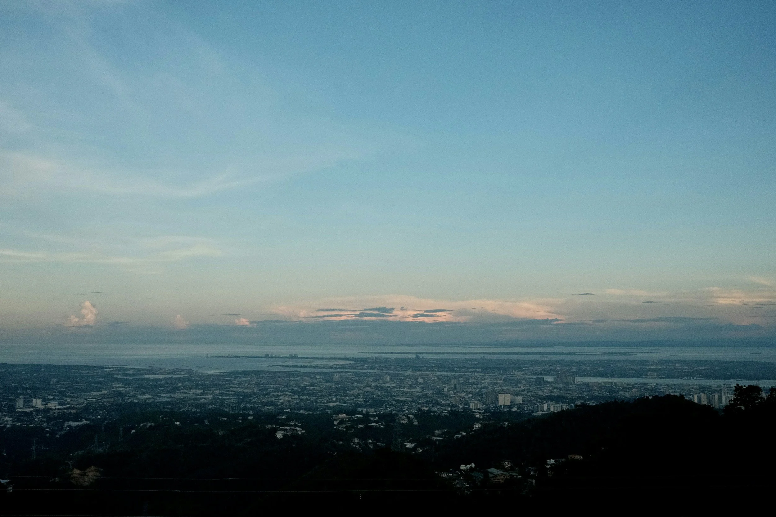 A wide elevated view looks over a city and distant water under layered clouds, the frame holding scale, haze, and quiet atmosphere.