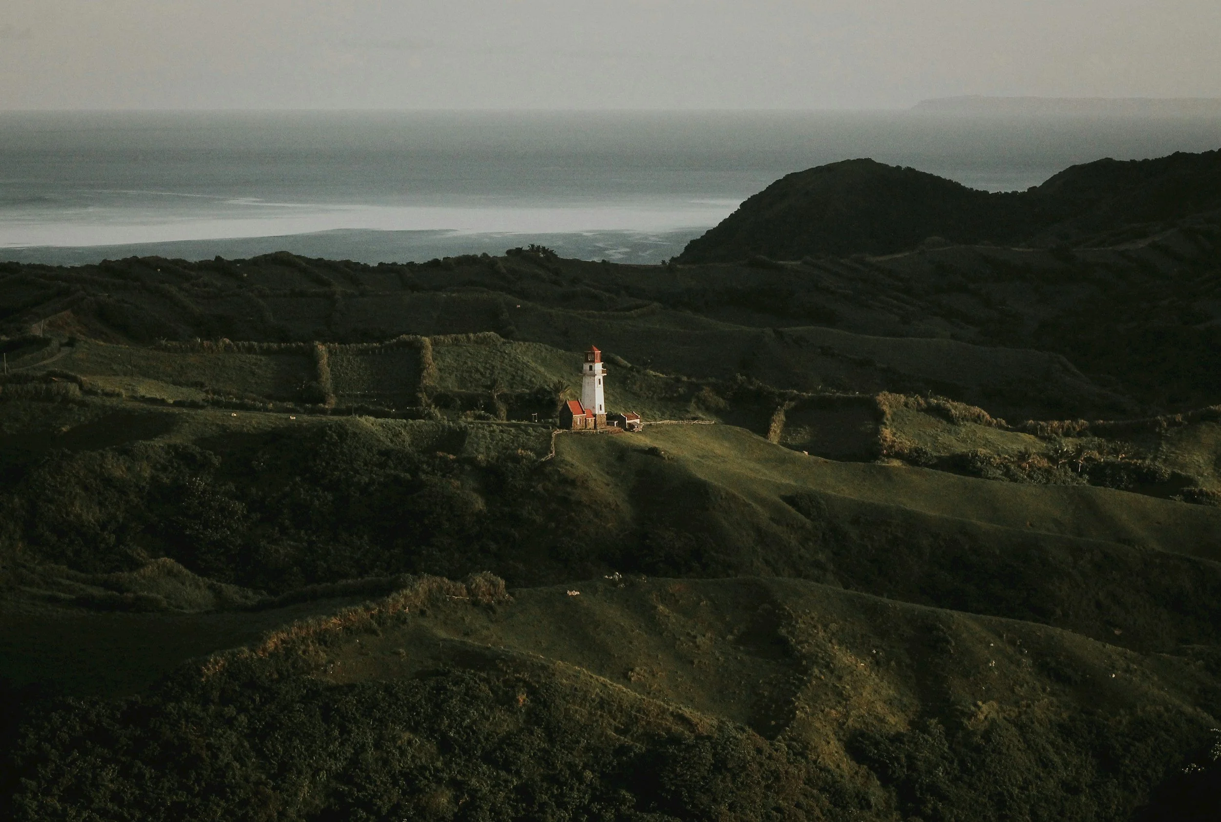 A solitary lighthouse rests among layered hills while low haze drifts across the scene, held like a cinematic prenup film still.