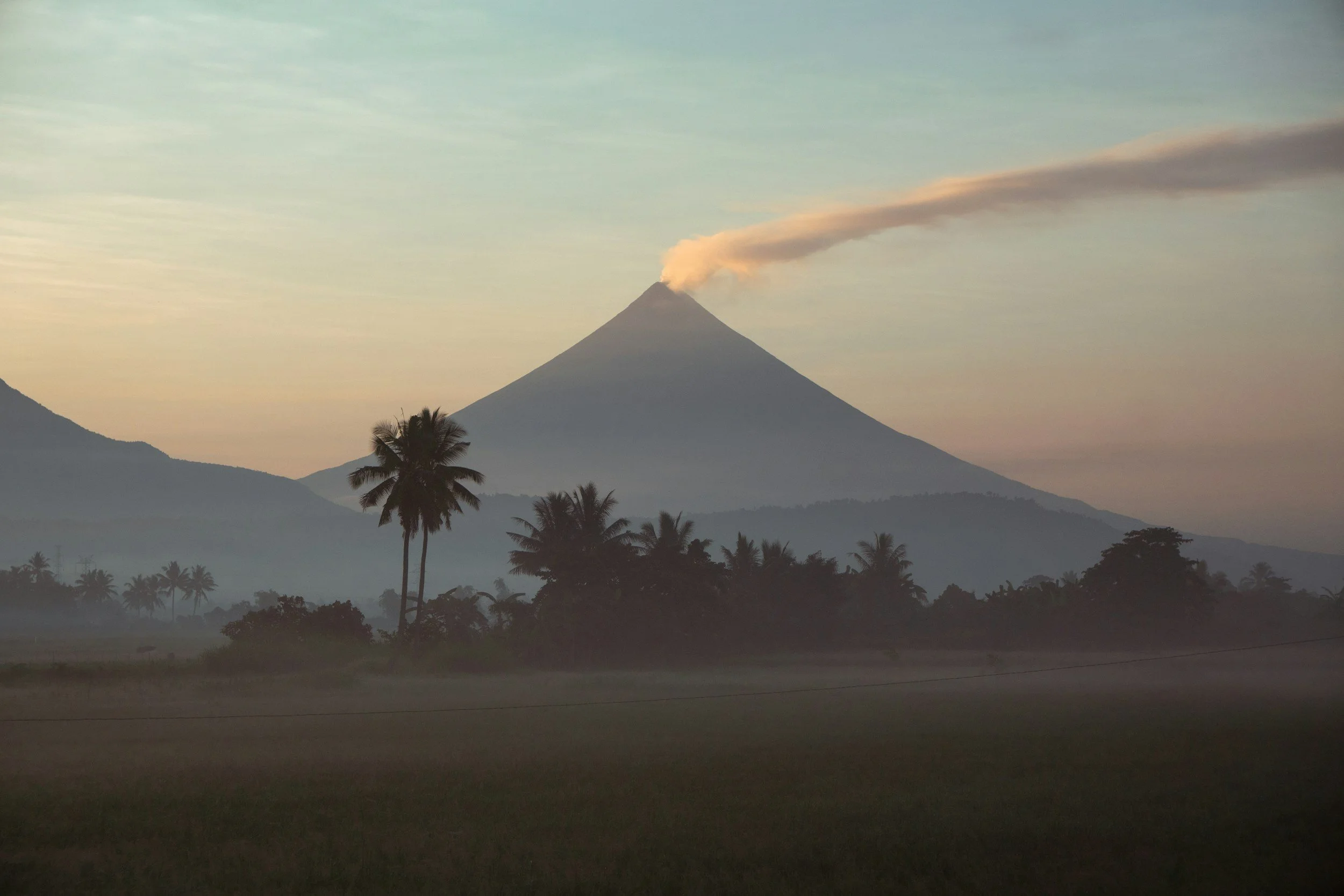 A distant mountain releases a thin plume into the sky while palm silhouettes stand still below shifting light.
