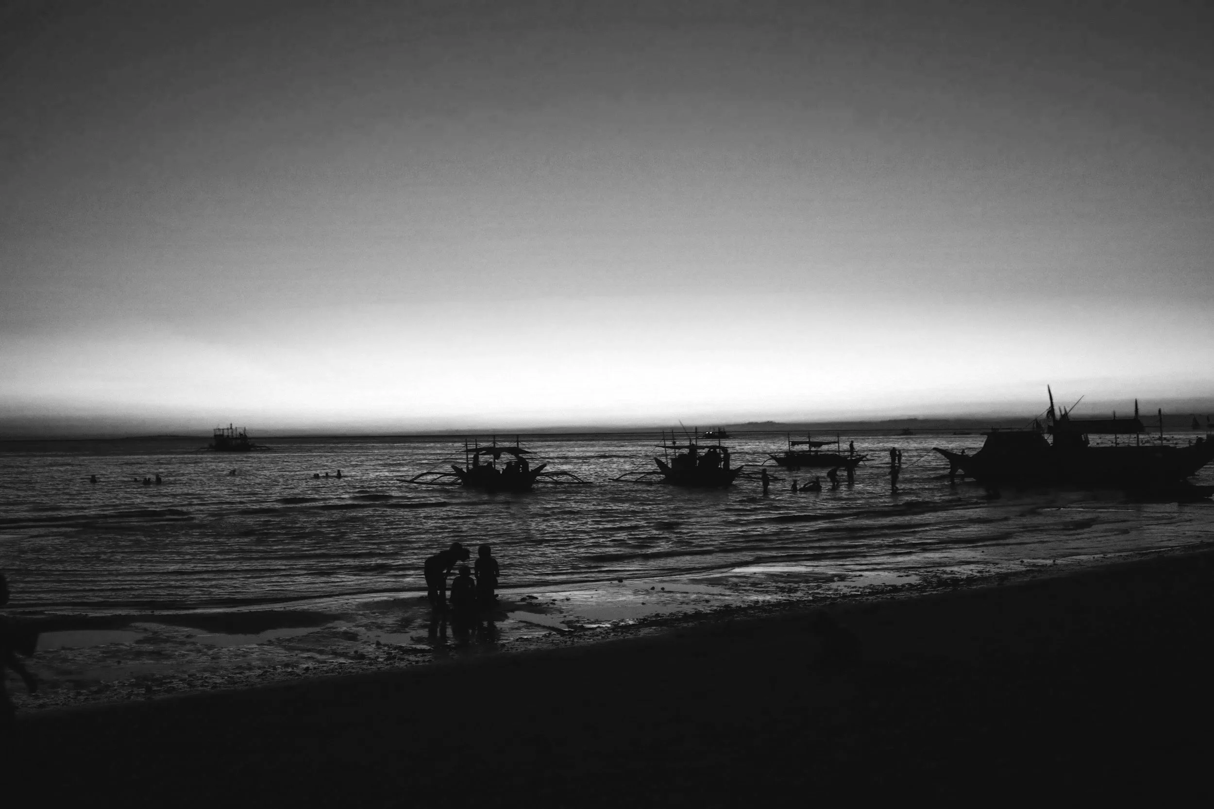 Small boats rest offshore at dusk while a couple stands near the waterline, framed quietly by the horizon and fading light.
