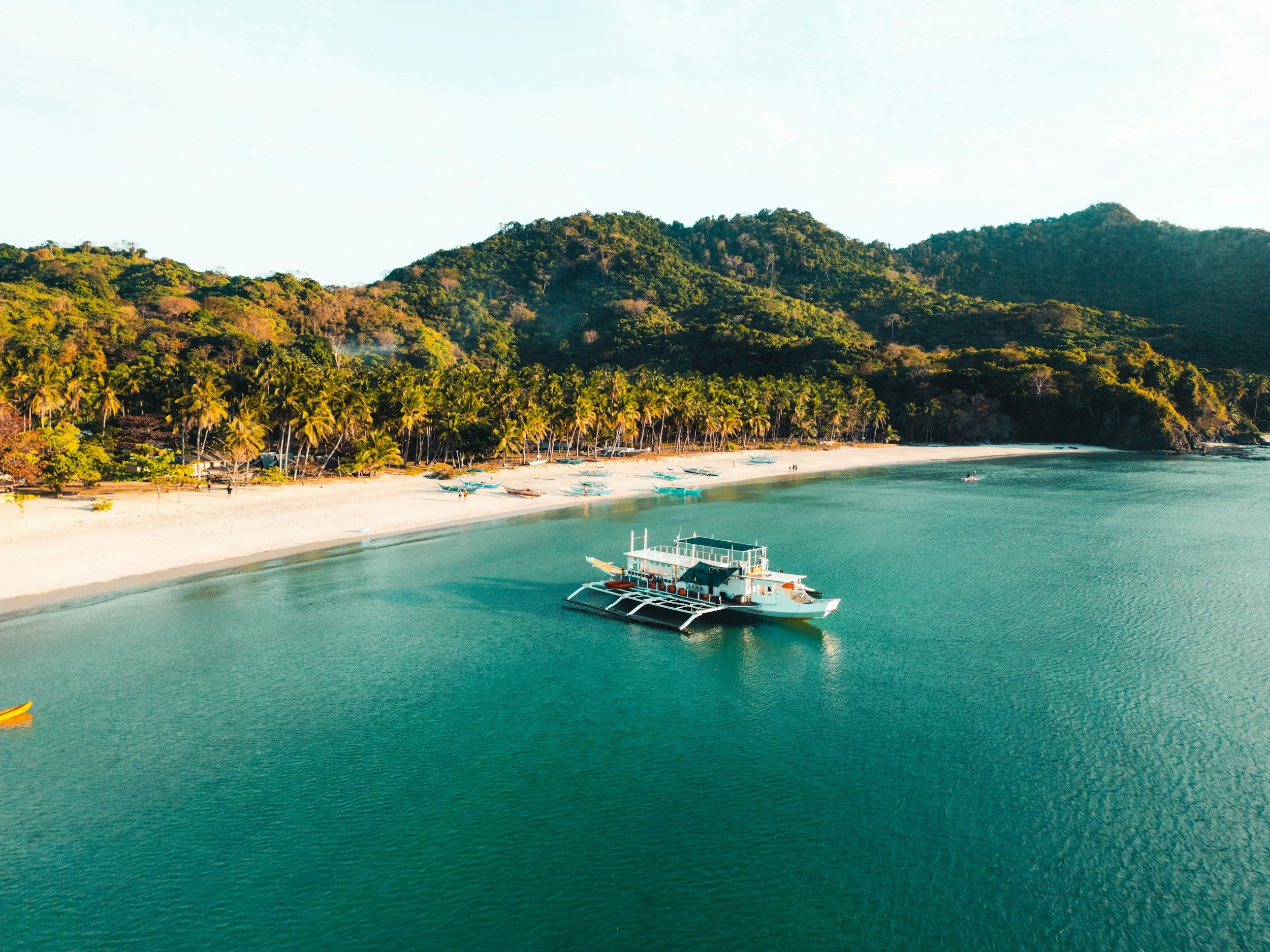 A small boat floats offshore near a curved beach while calm water holds steady beneath distant hills in a cinematic prenup film frame.
