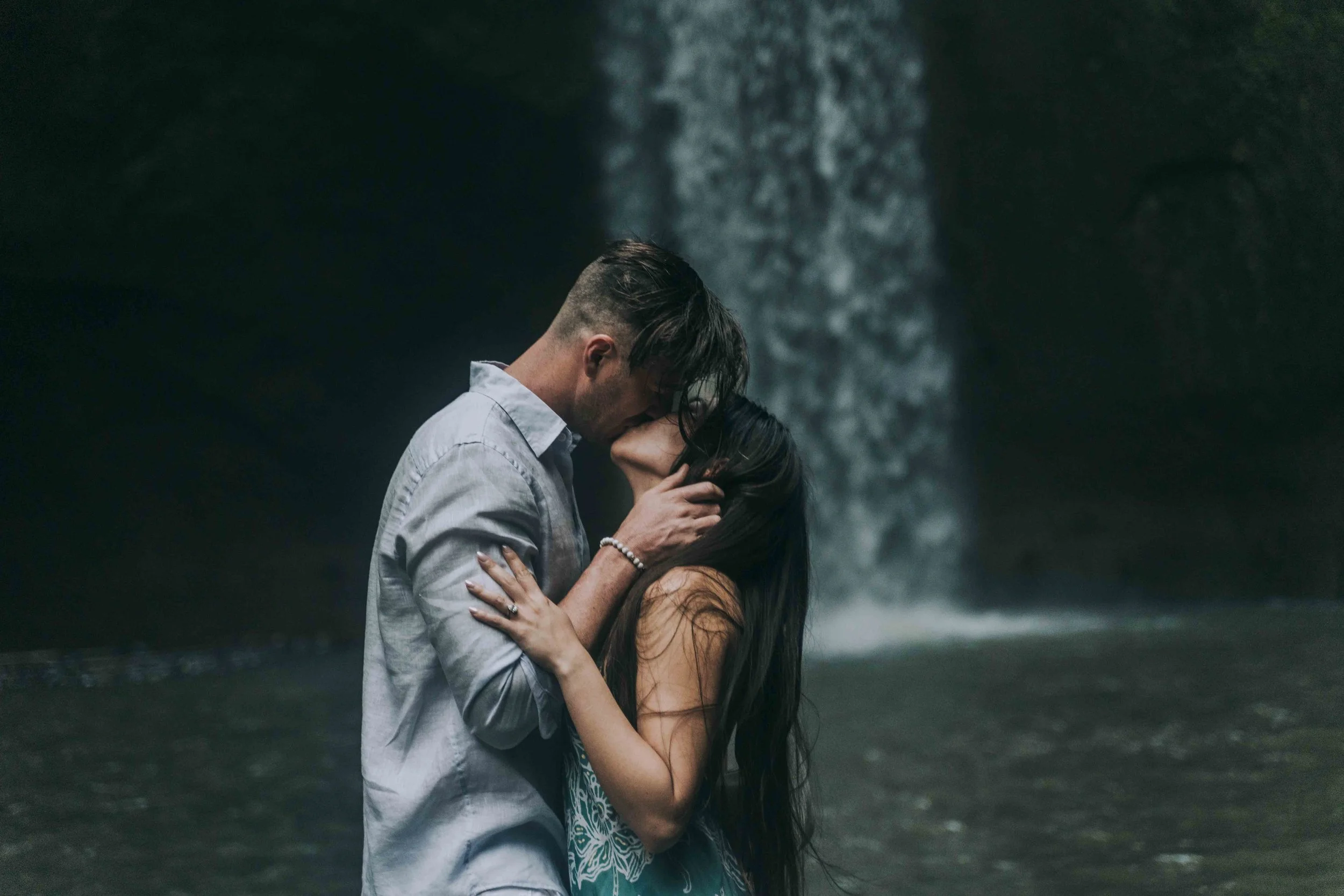 A couple embraces beneath falling water, bodies pressed close as movement blurs into a cinematic prenup film moment.
