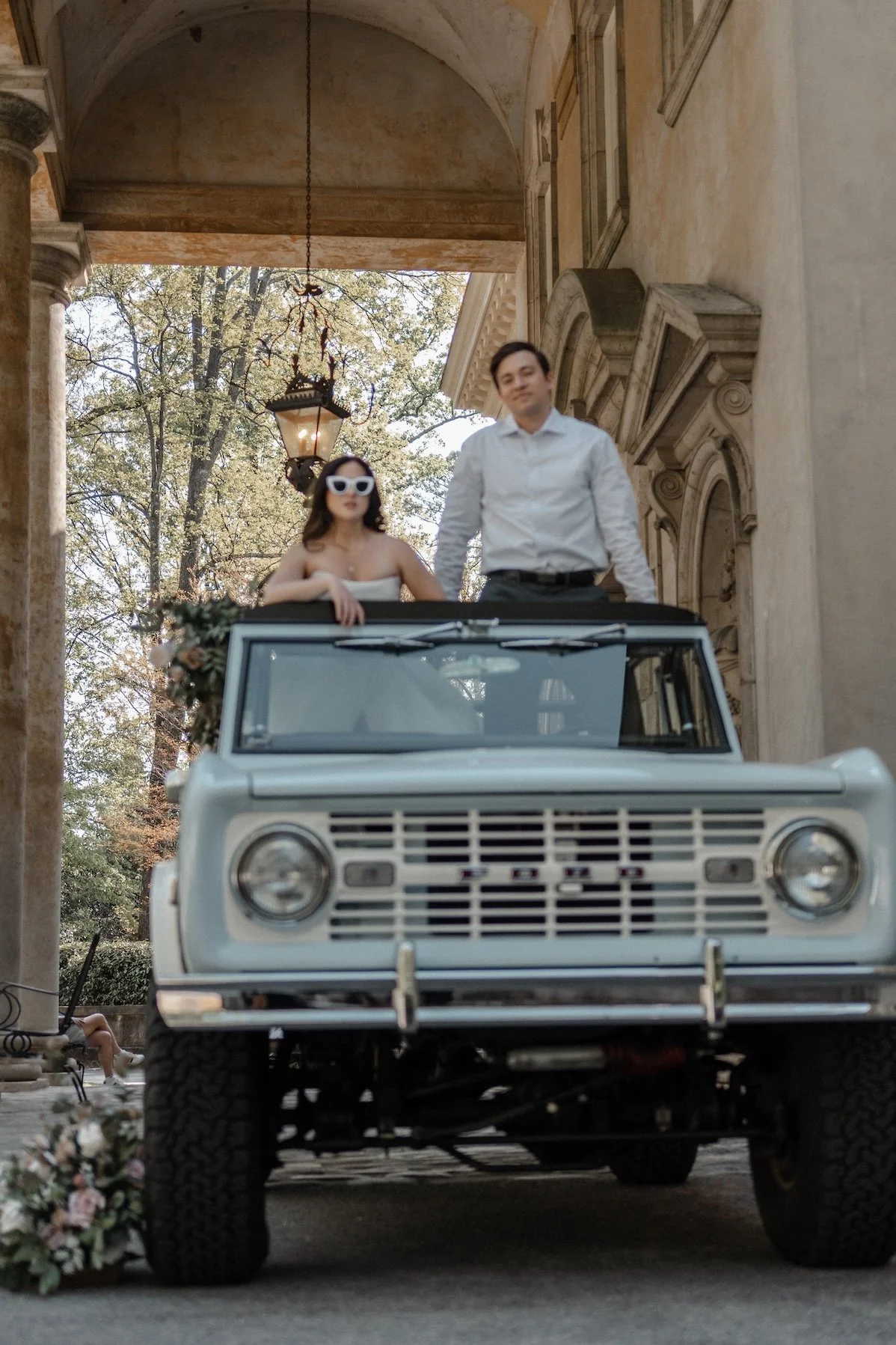 A groom standing beside a vintage car with a bride seated above, the scene playful and romantic.