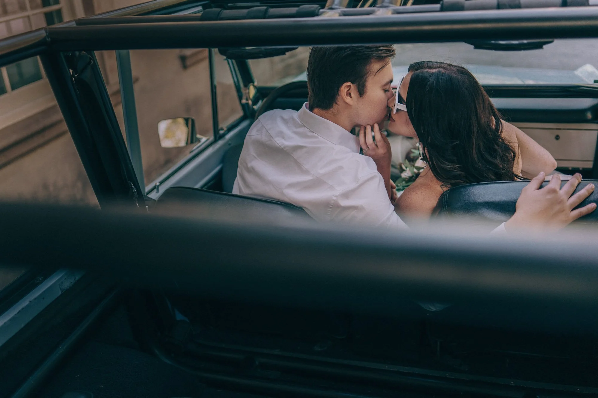 A couple leans toward each other inside a parked vehicle, framed through the window by a Fragmented Memories prenup photographer watching from outside.