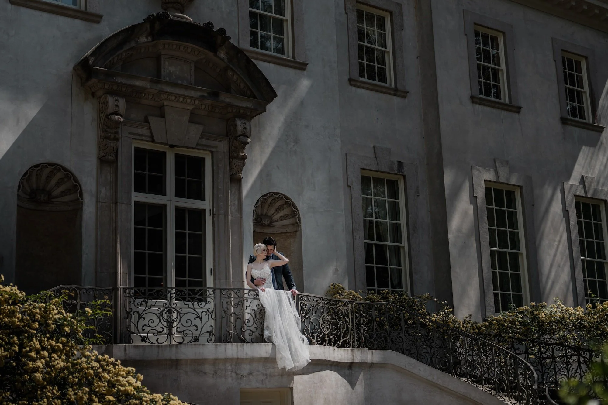 Bride and groom embrace on a terrace balcony, shadows and columns creating an atmospheric editorial frame by Caz Isaiah