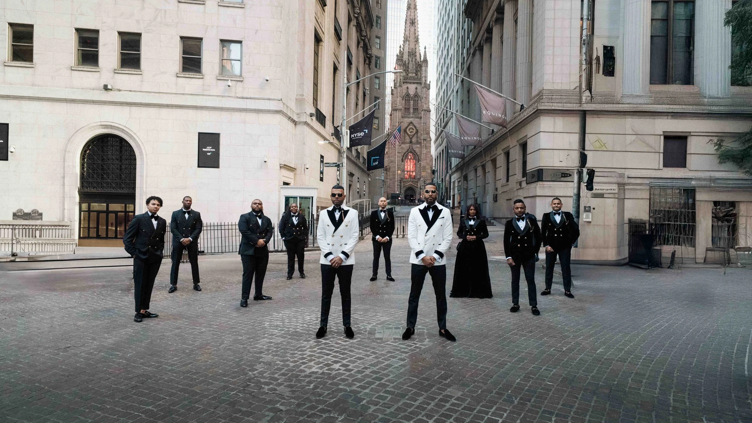 Groom and groomsmen stand in formation on cobblestone street, formal black and white attire beneath tall buildings, cinematic and editorial