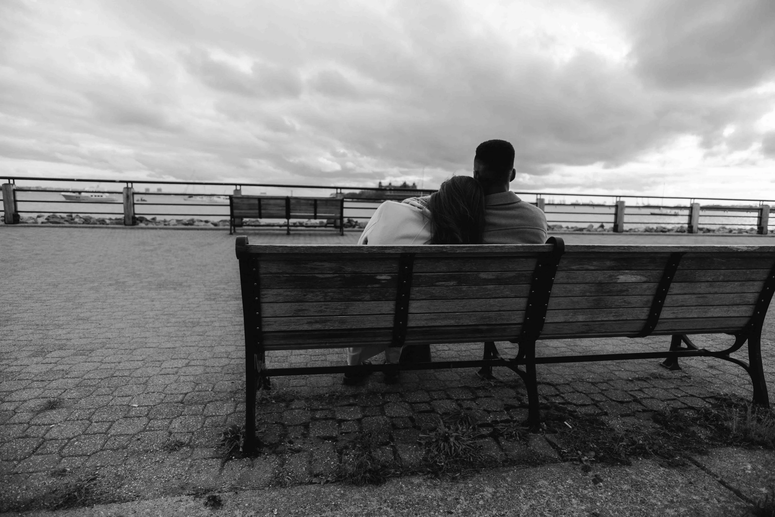 A couple sits close on a wooden bench facing open water, framed quietly by a Fragmented Memories prenup photographer observing distance and stillness.