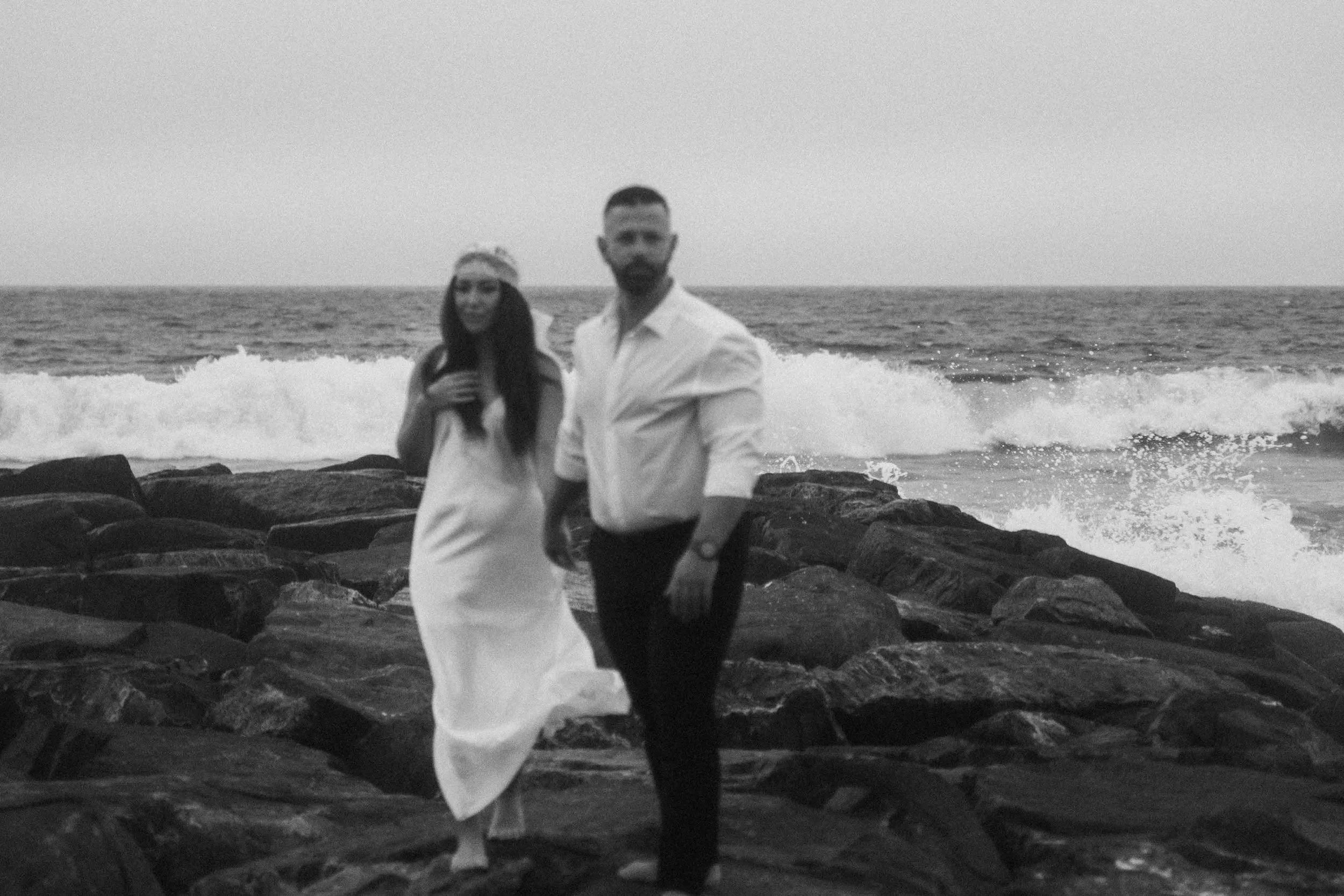 A man and woman walk across dark rocks near crashing waves, bodies moving forward together like a slowed cinematic couples film moment.