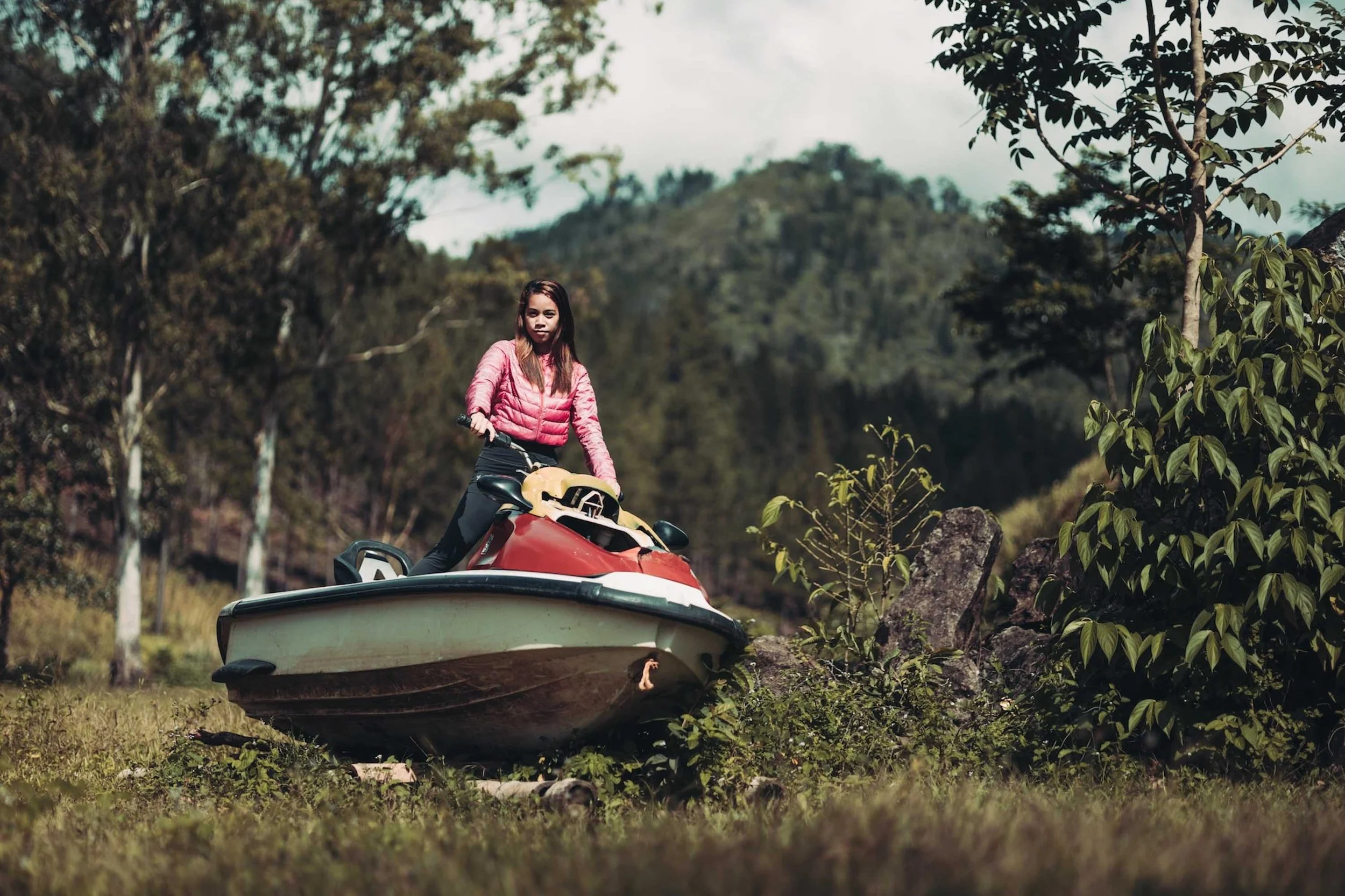 A woman beside a small boat on land, hands resting against it, surrounded by jungle trees and soft golden light.