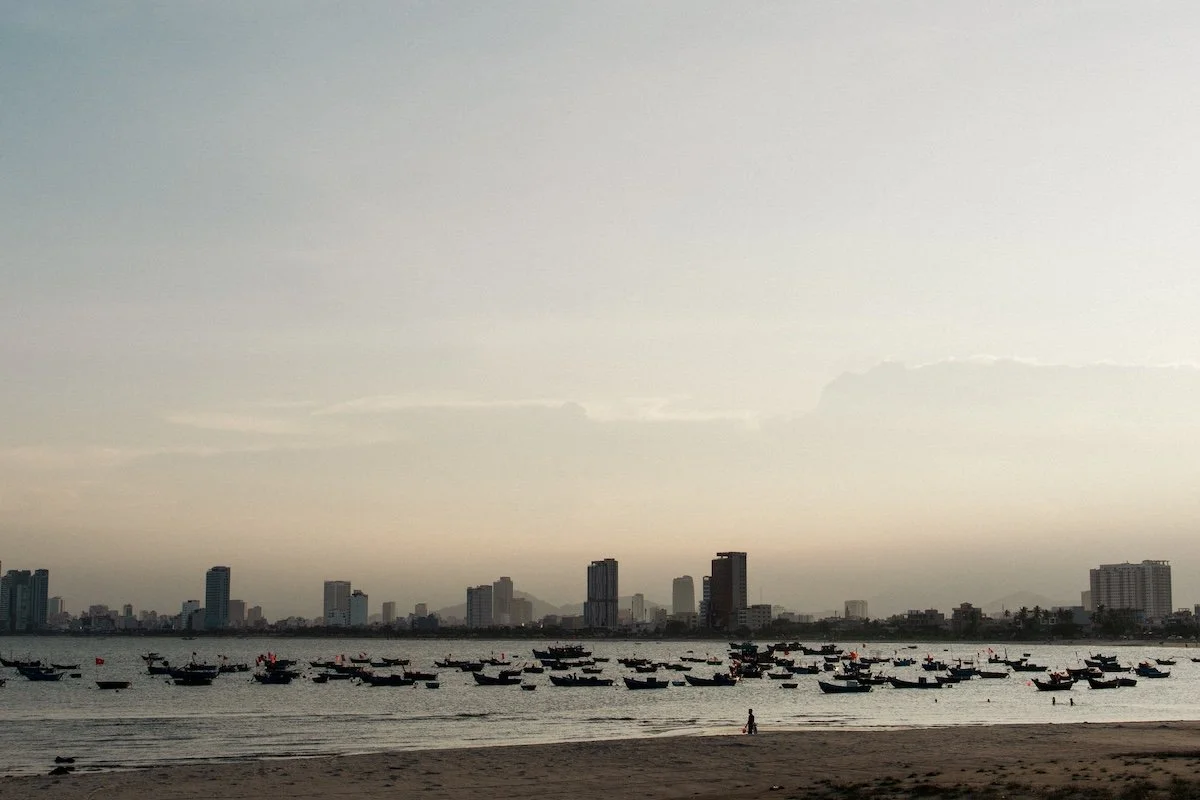 The city skyline fading into dusk, boats scattered across calm water, cinematic coastal scene of Da Nang