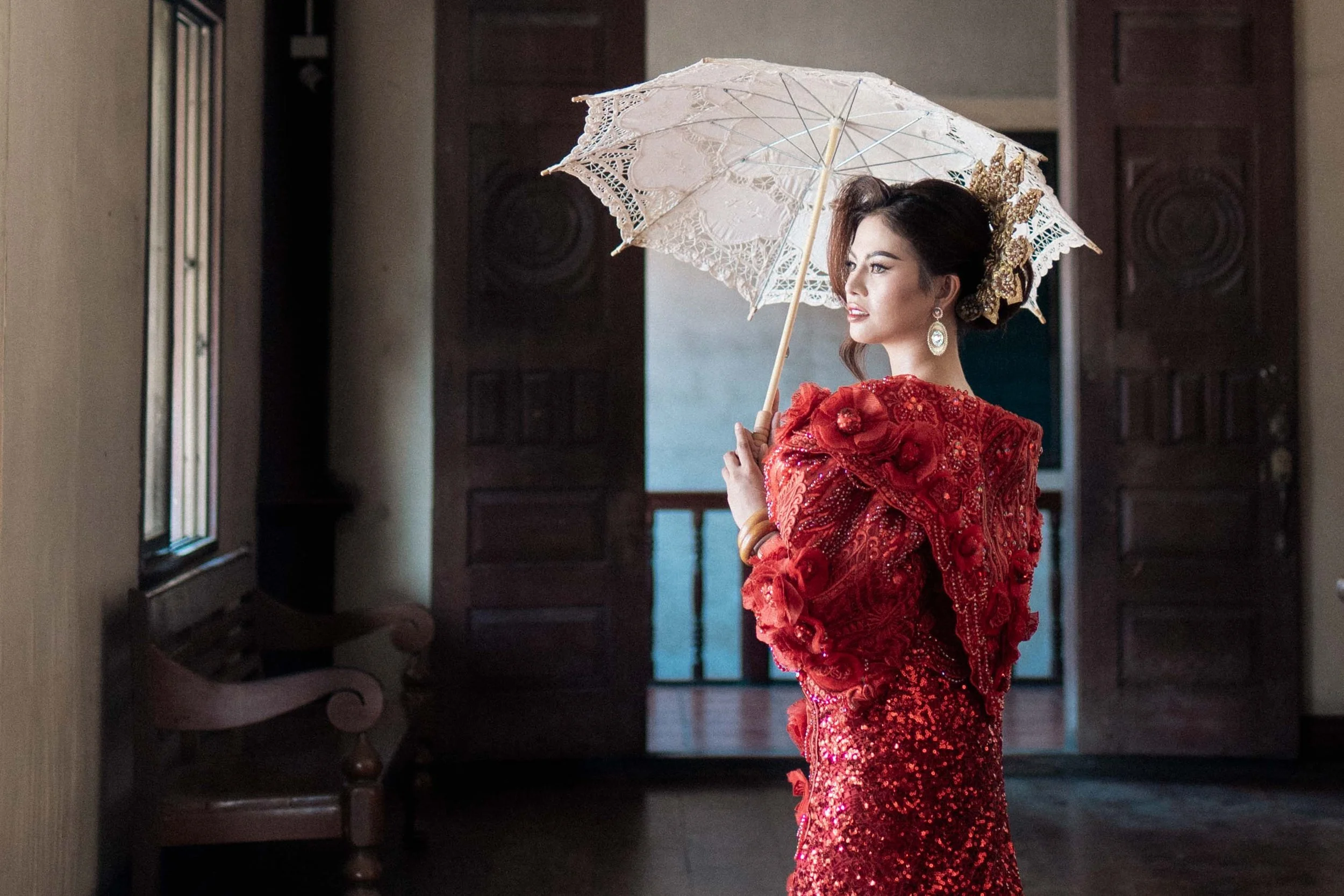 A bride in a red dress holds an umbrella in soft window light, cinematic portraits by Caz Isaiah in the Philippines.