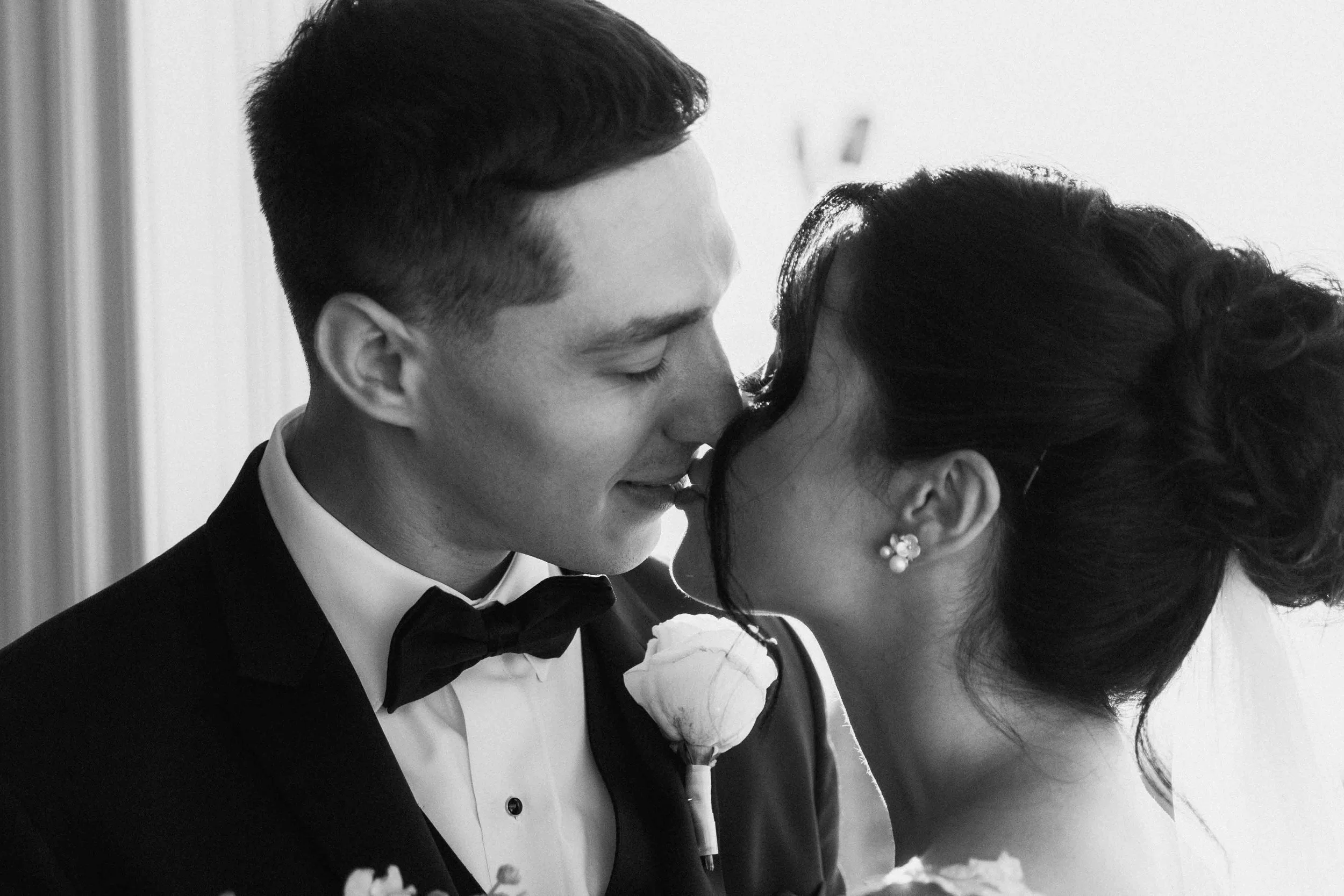 In close frame, a bride and groom touch noses and kiss softly, photographed in black and white by Caz Isaiah.