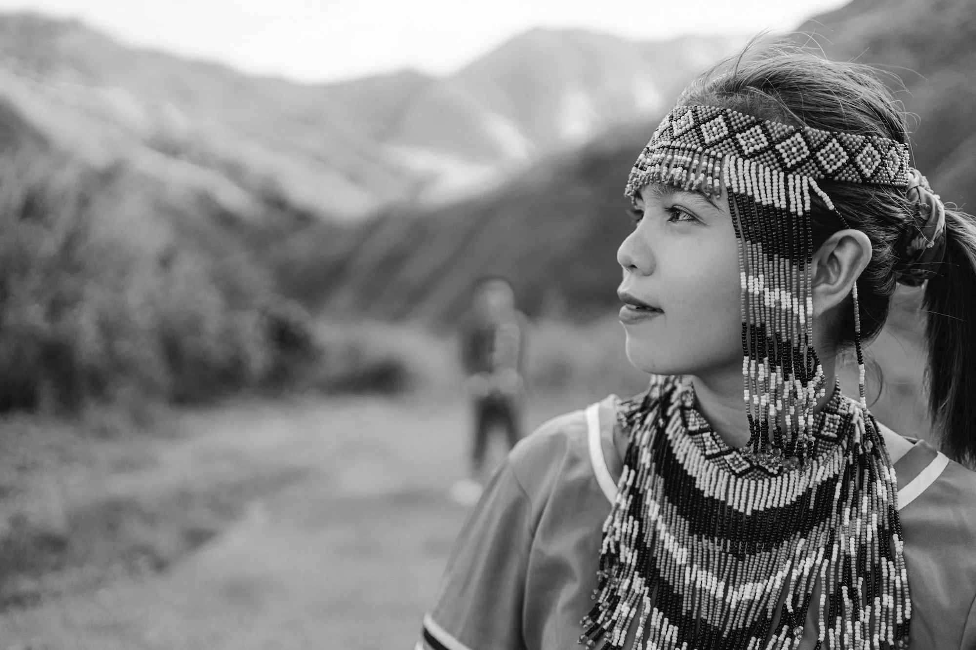 A woman wearing a beaded headpiece looks to the side on a mountain path as a figure stands blurred behind her, photographed by a Fragmented Memories couples photographer.