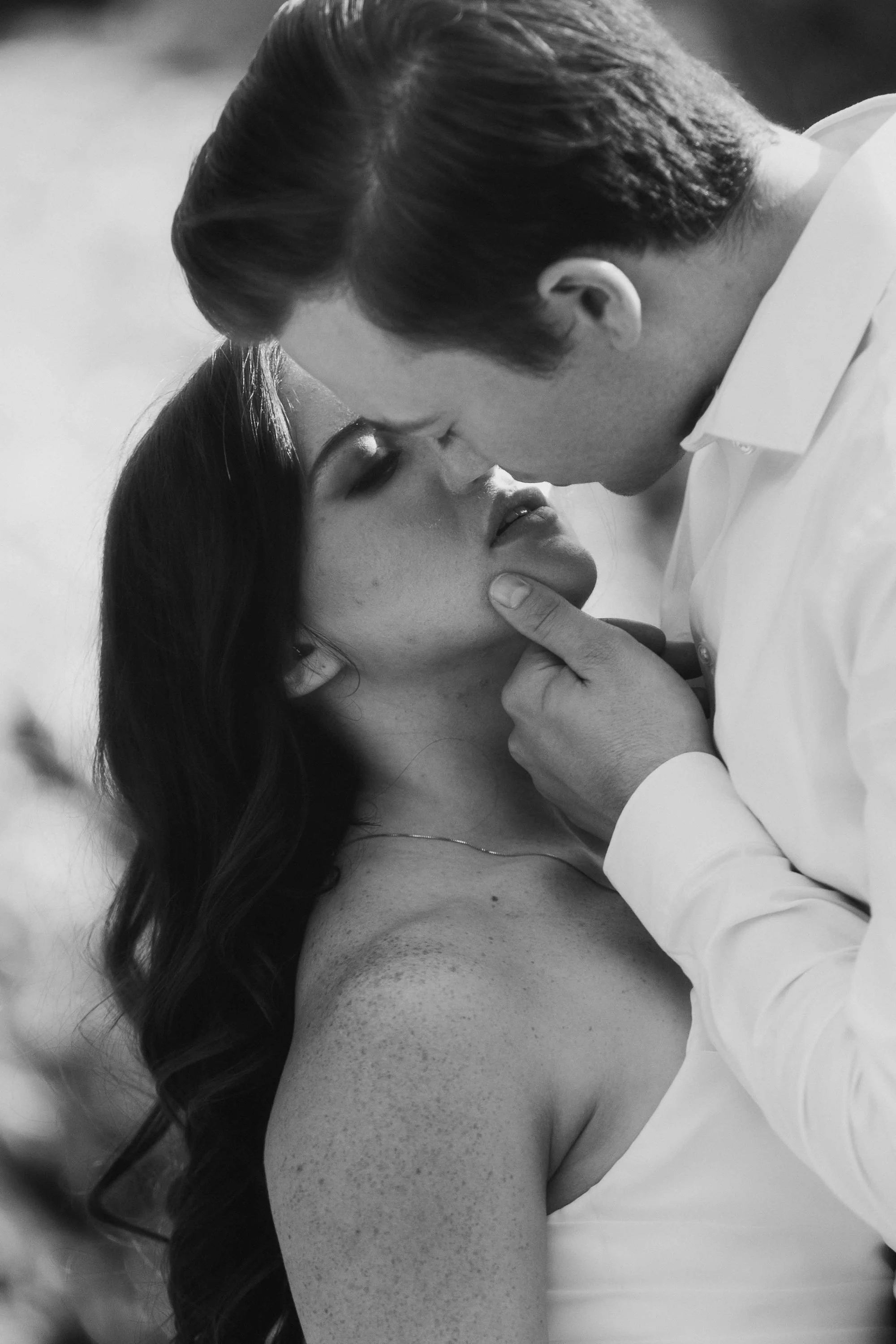 Black-and-white editorial kiss — groom gently holding his bride in close embrace.