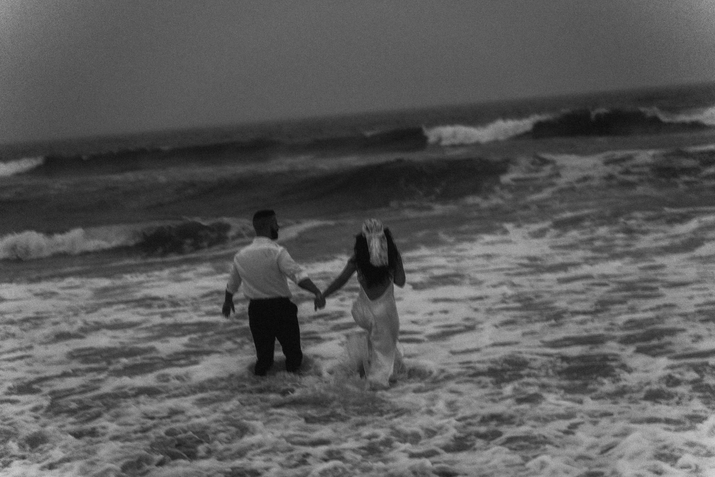 A couple walks hand in hand through breaking waves, clothing soaked as surf rises around them, captured by a Fragmented Memories couples photographer.