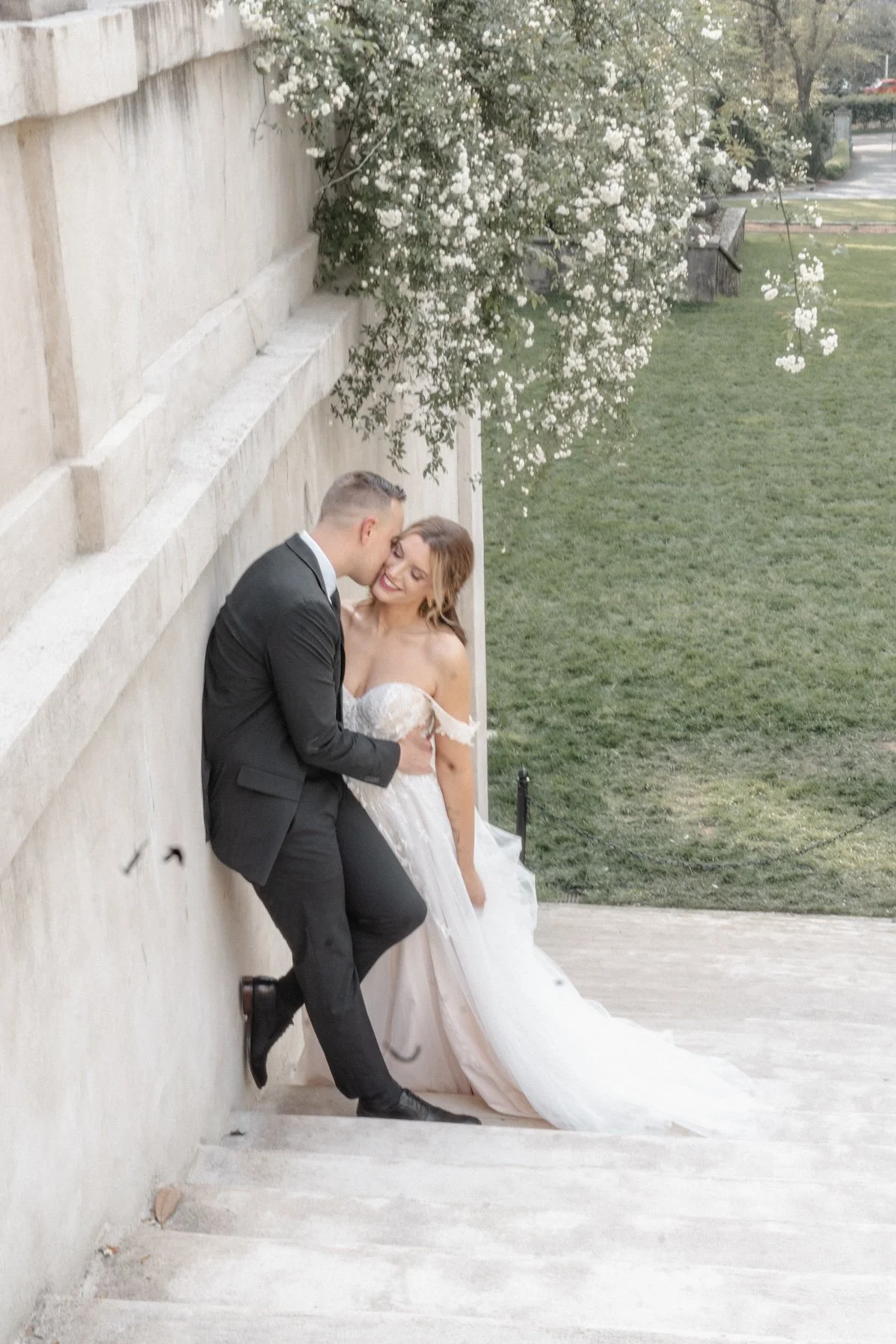 A couple leaning together against stone walls, framed in elegant wedding attire.
