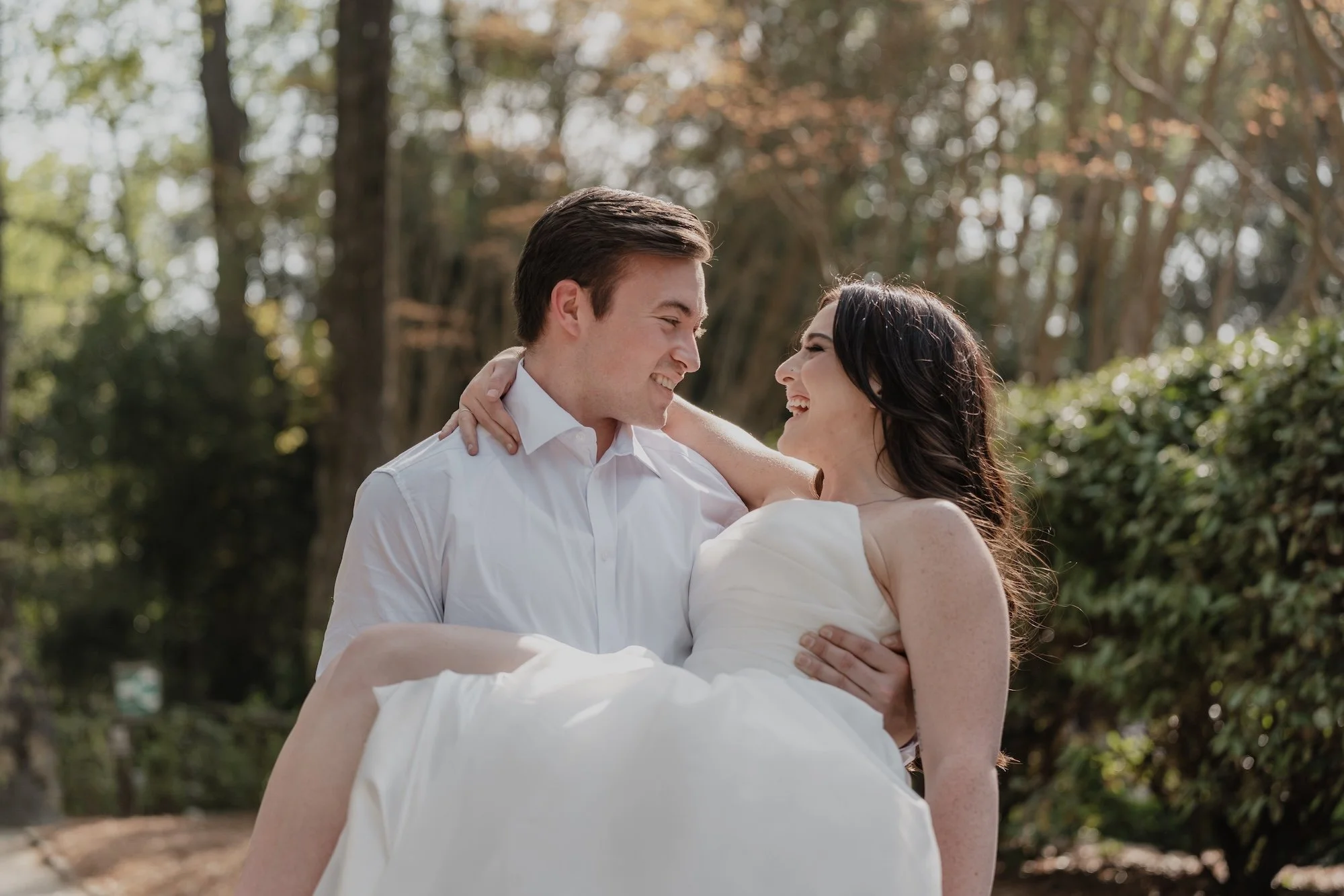 A groom lifts his bride into his arms, sunlight pouring through the trees, their laughter soft and unposed — wedding photography in Vietnam