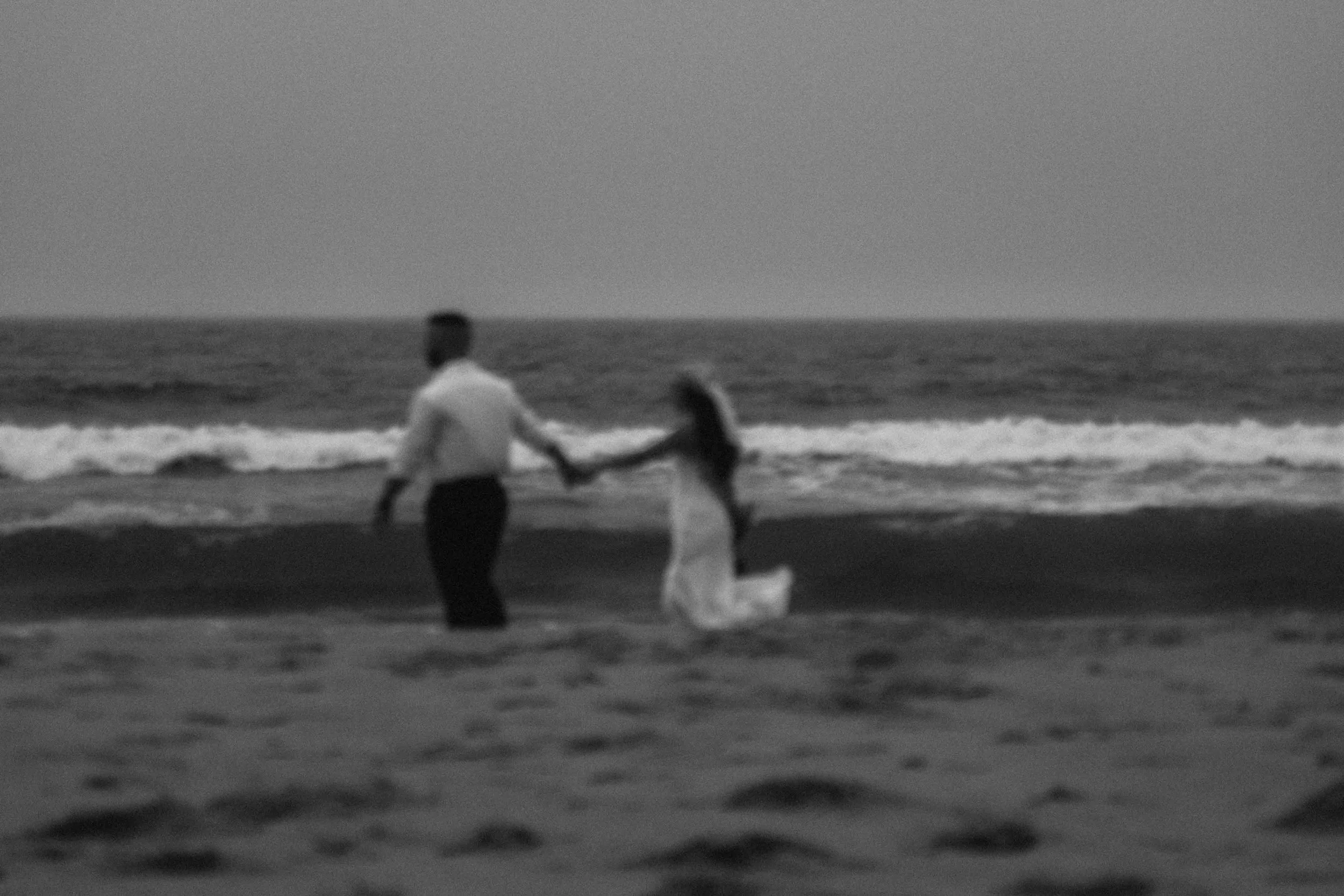 A couple walks hand in hand toward the shoreline as waves rise behind them, captured with the quiet tension of a cinematic couples film.