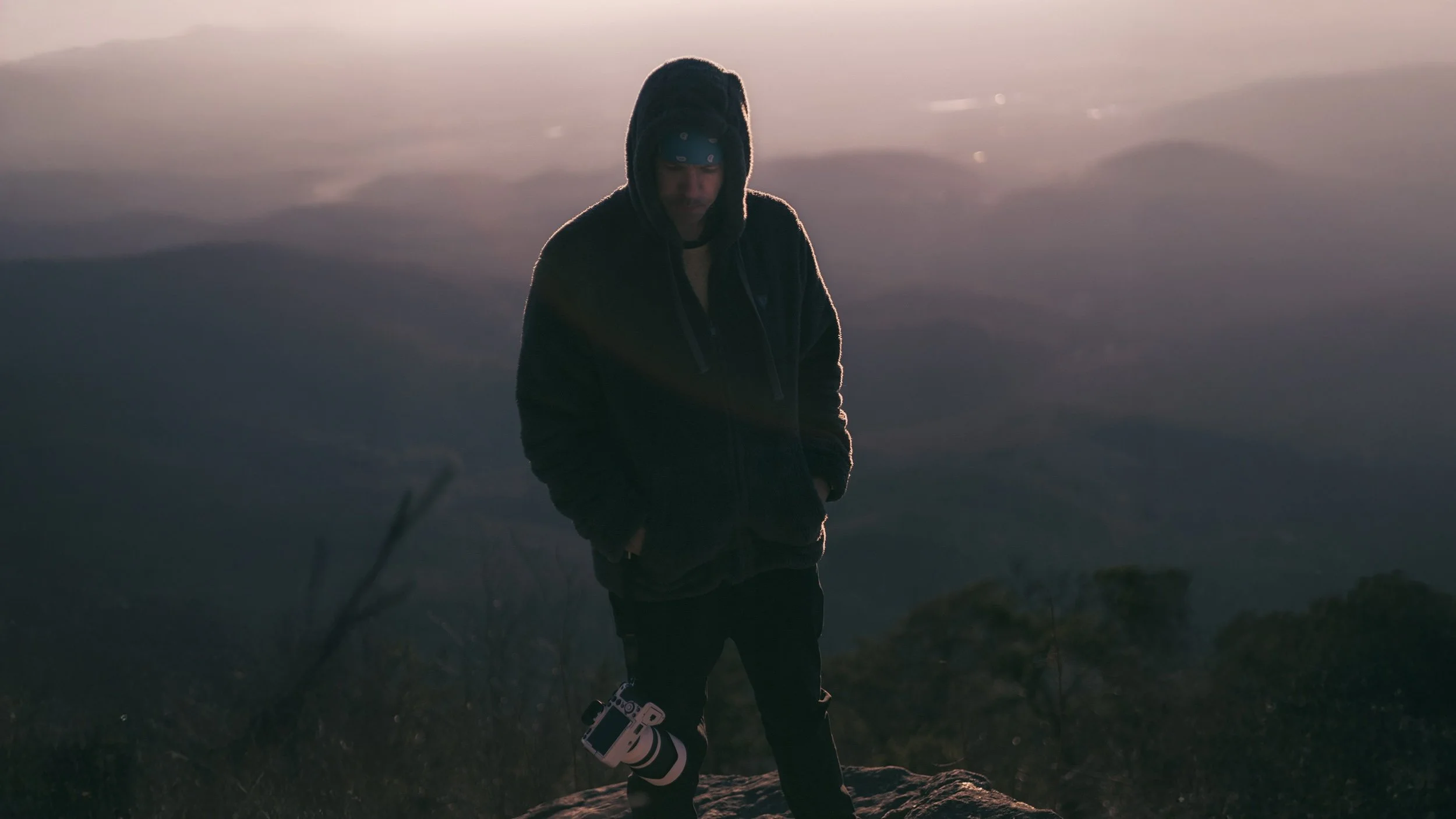 A hooded figure stands alone on a mountain ridge at dusk, camera hanging at his side as layered hills fade into low light.