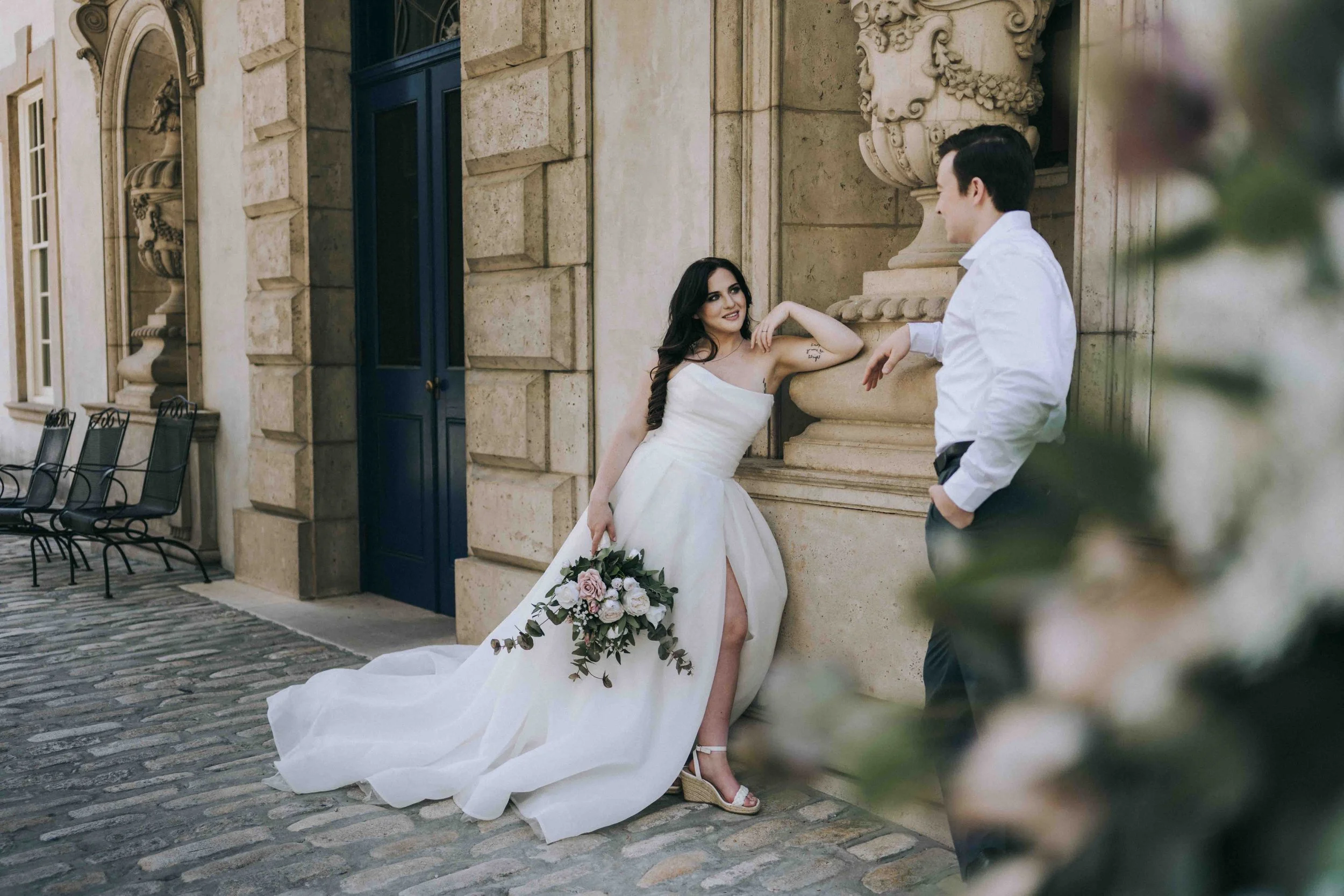 Bride leans against a stone wall holding a floral bouquet while the groom stands nearby, both framed in soft golden light. Captured by Bali-based wedding photographer Caz Isaiah, inspired by the coastal elegance of Uluwatu.