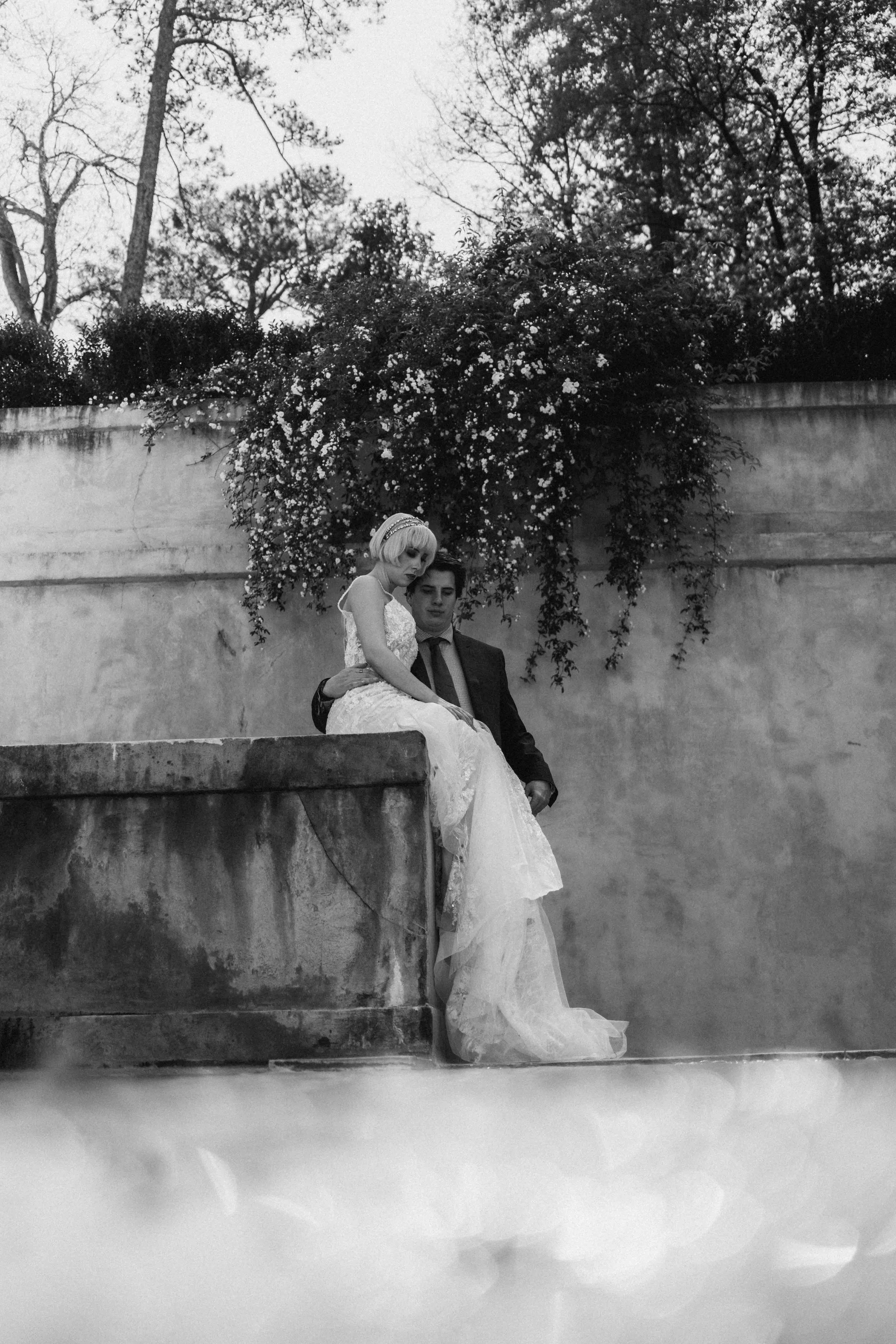 Couple seated in quiet embrace against a stone wall, captured by Caz Isaiah in a timeless black-and-white editorial frame inspired by Oman’s serene architecture.