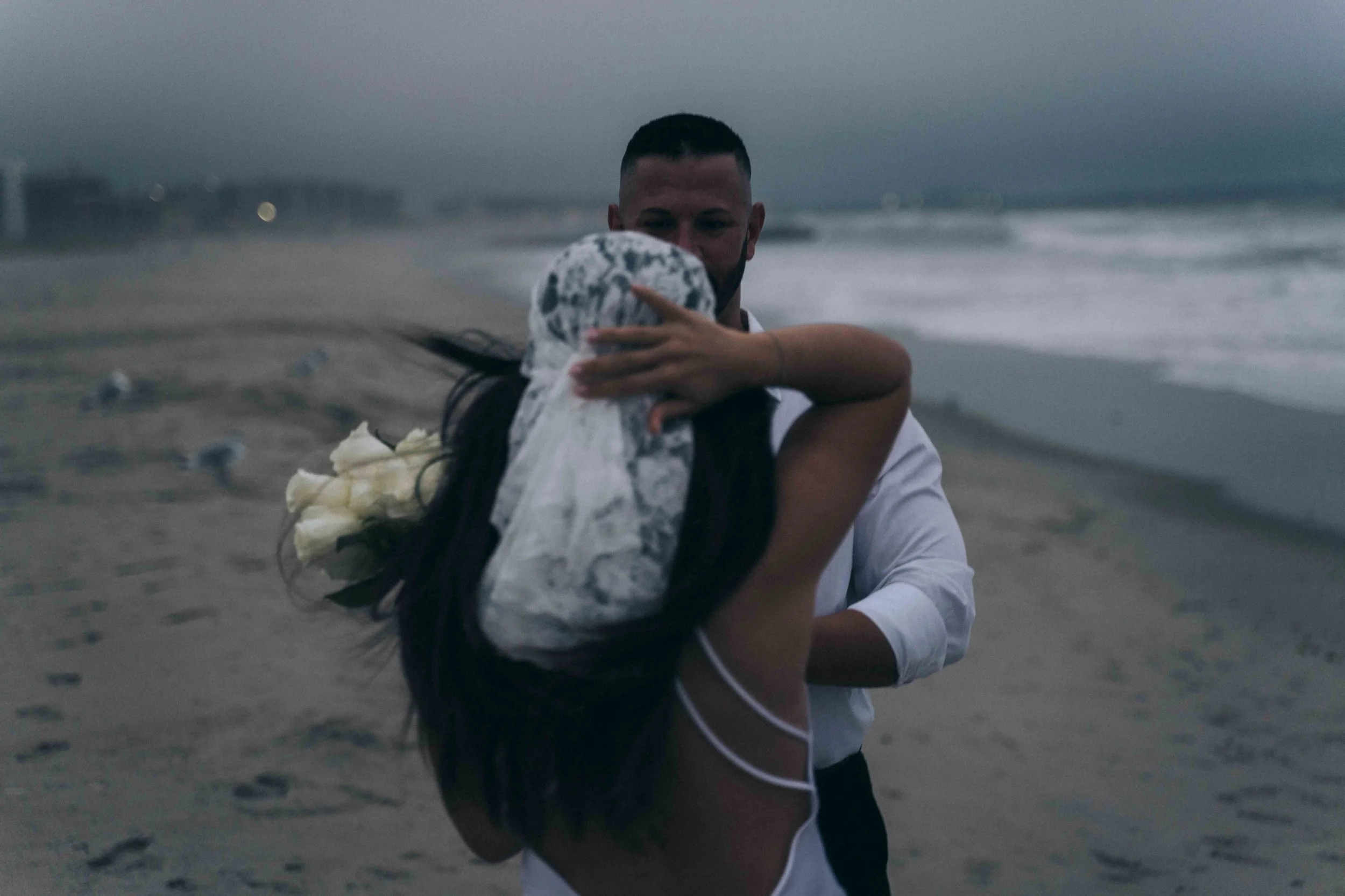 Two figures embrace near the shoreline, bodies turned toward each other as dark water moves behind them under a muted sky.