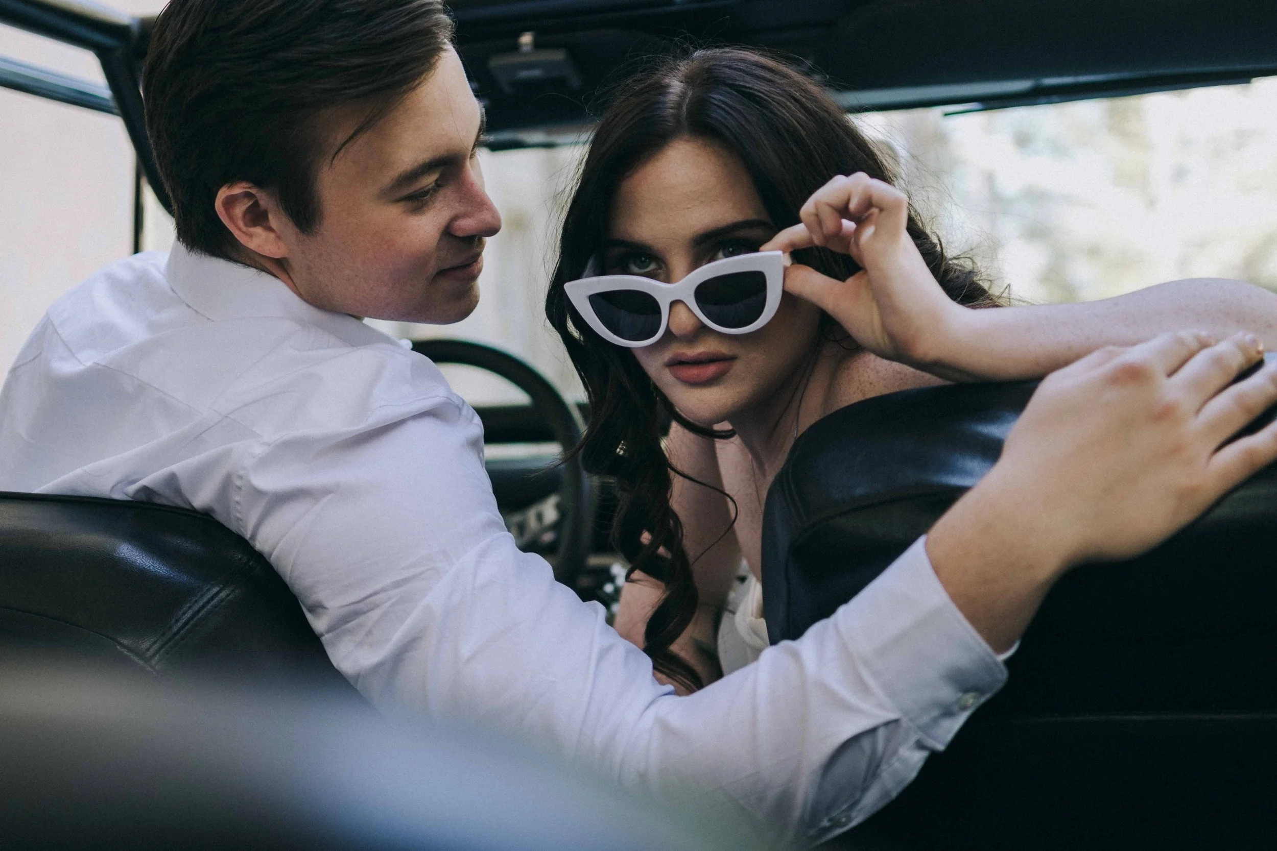 A woman turns toward the camera inside a vehicle, adjusting her sunglasses while a man leans close beside her in quiet proximity.