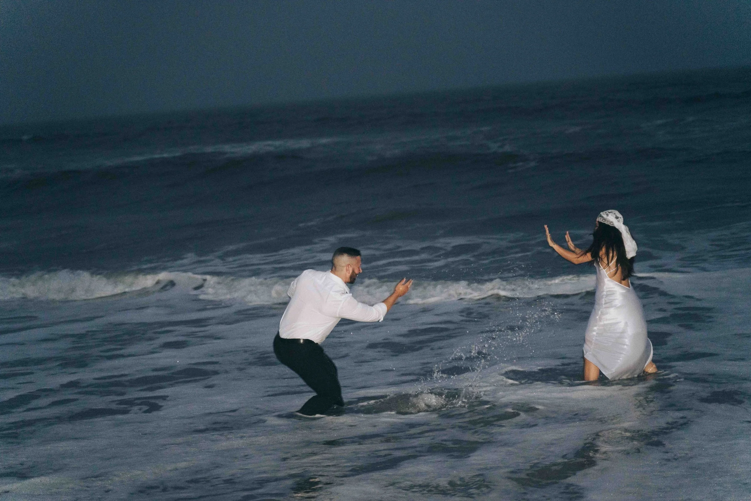 A couple splashes toward each other in shallow surf, arms raised mid motion, unfolding as a cinematic prenup film against rolling waves.