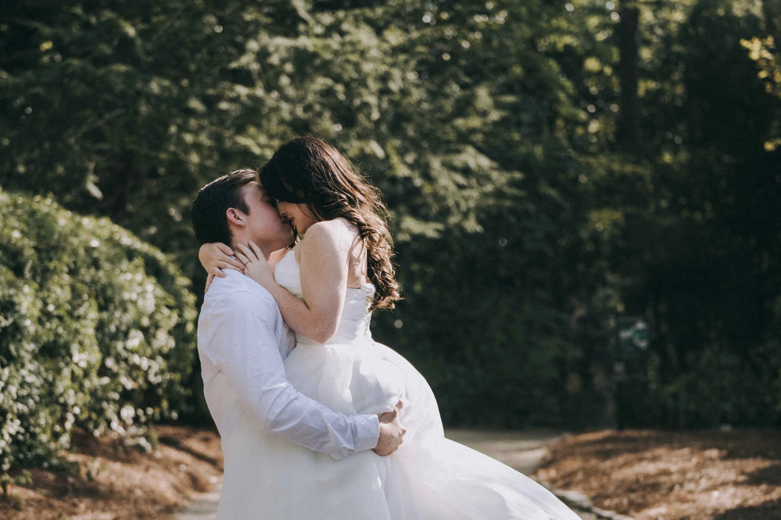 A couple embraces on a shaded path, bodies pressed together as filtered light settles softly through surrounding trees.