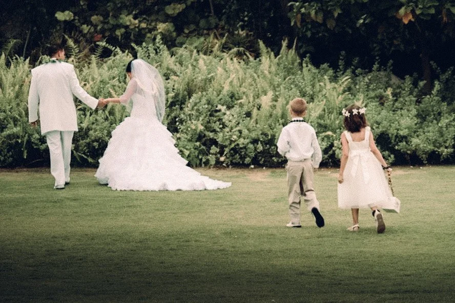 Bride walks hand in hand with children across the grass, veil trailing softly in the light