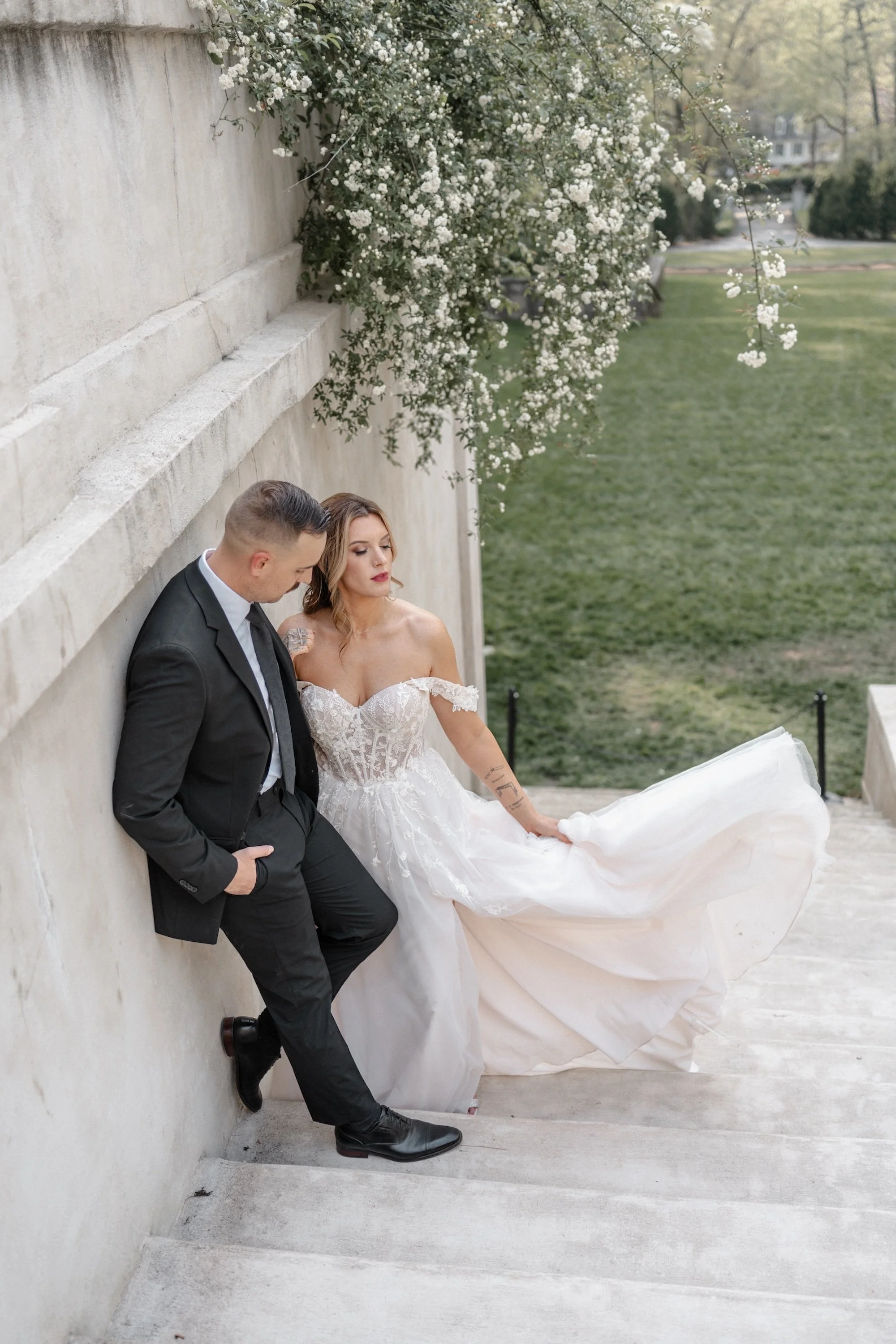 The couple leans close on marble steps, her dress spilling into soft folds as they share a private moment.