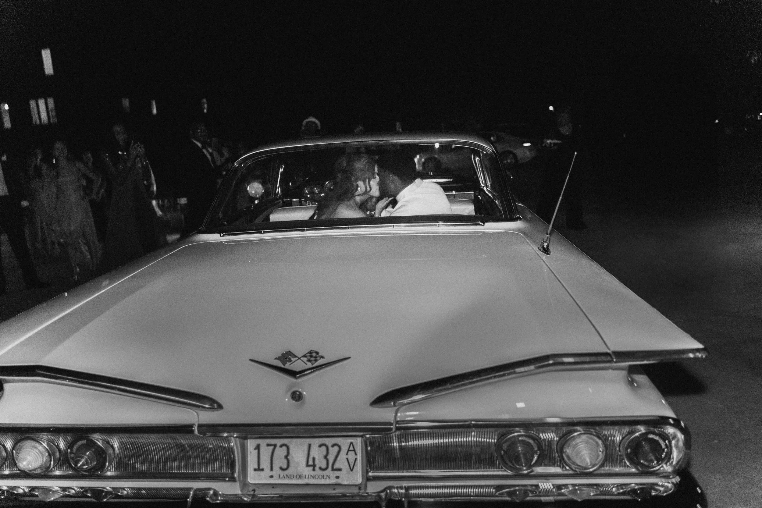 A newlywed couple drives away in a vintage car, headlights glinting through the night — cinematic moment by Caz Isaiah.