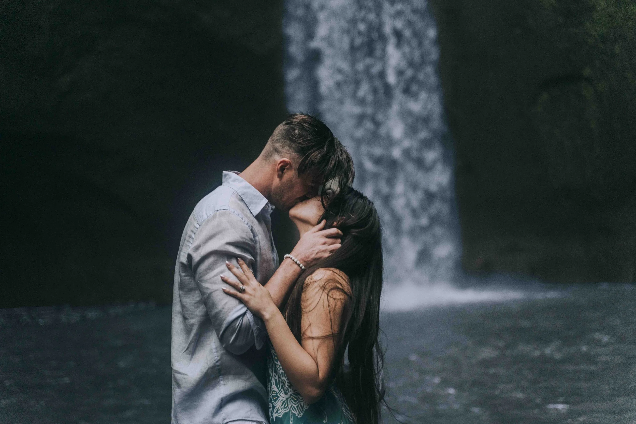 A couple stands close beneath a waterfall, lips touching, water and stone enclosing the frame.