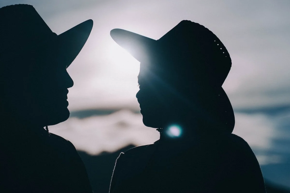 Two figures in wide-brim hats leaning close, their silhouettes glowing in backlight in the mountains