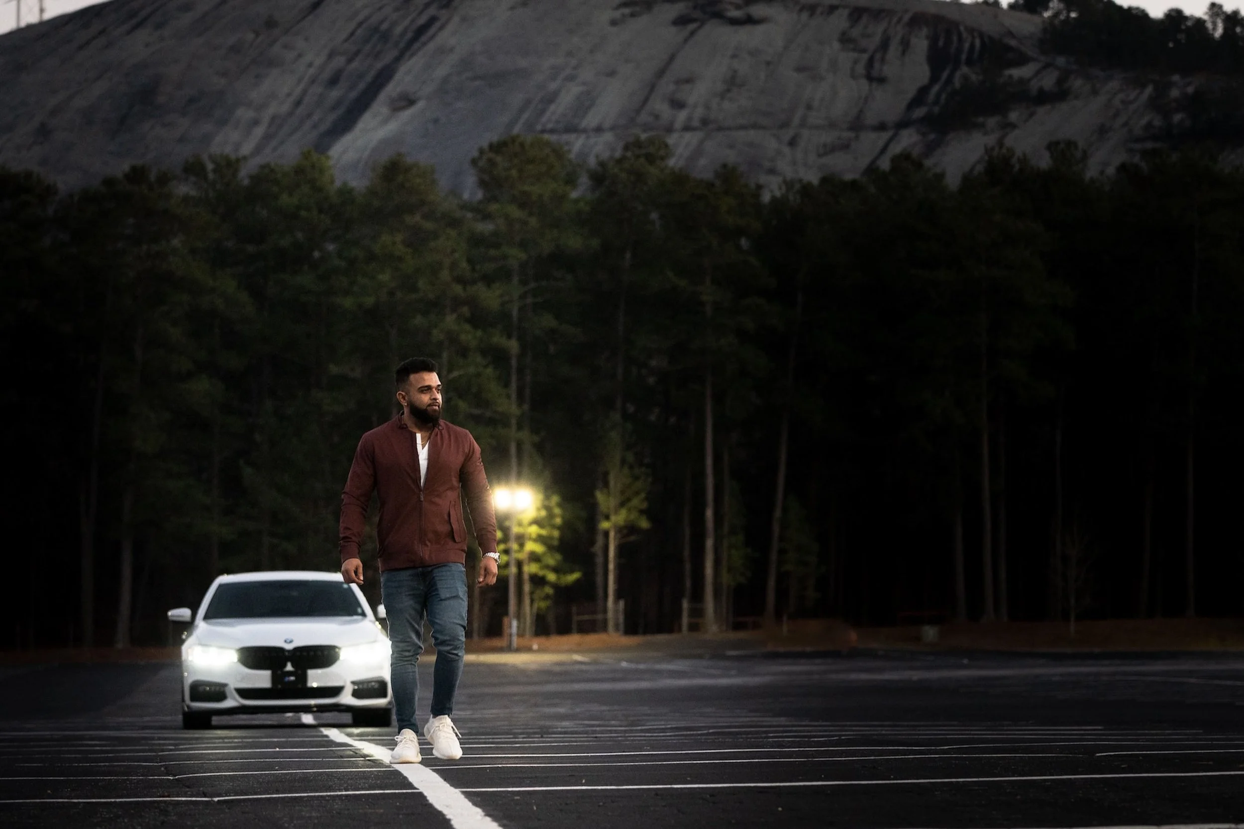 A man walks along a rain-dark road under a streetlight, mist rising around a parked car.