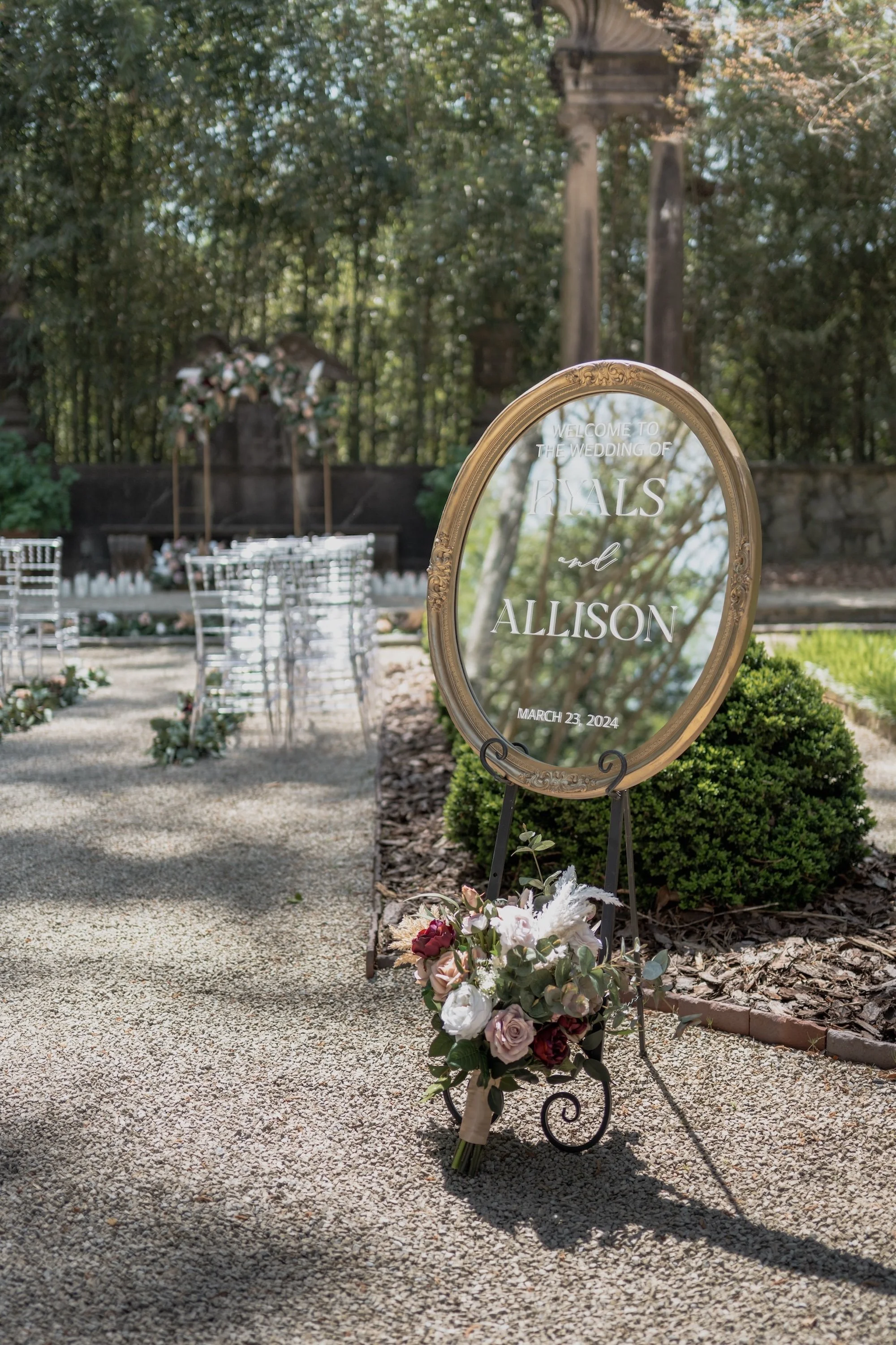 Ceremony sign set among greenery, reflecting the couple’s names in a polished gold frame.