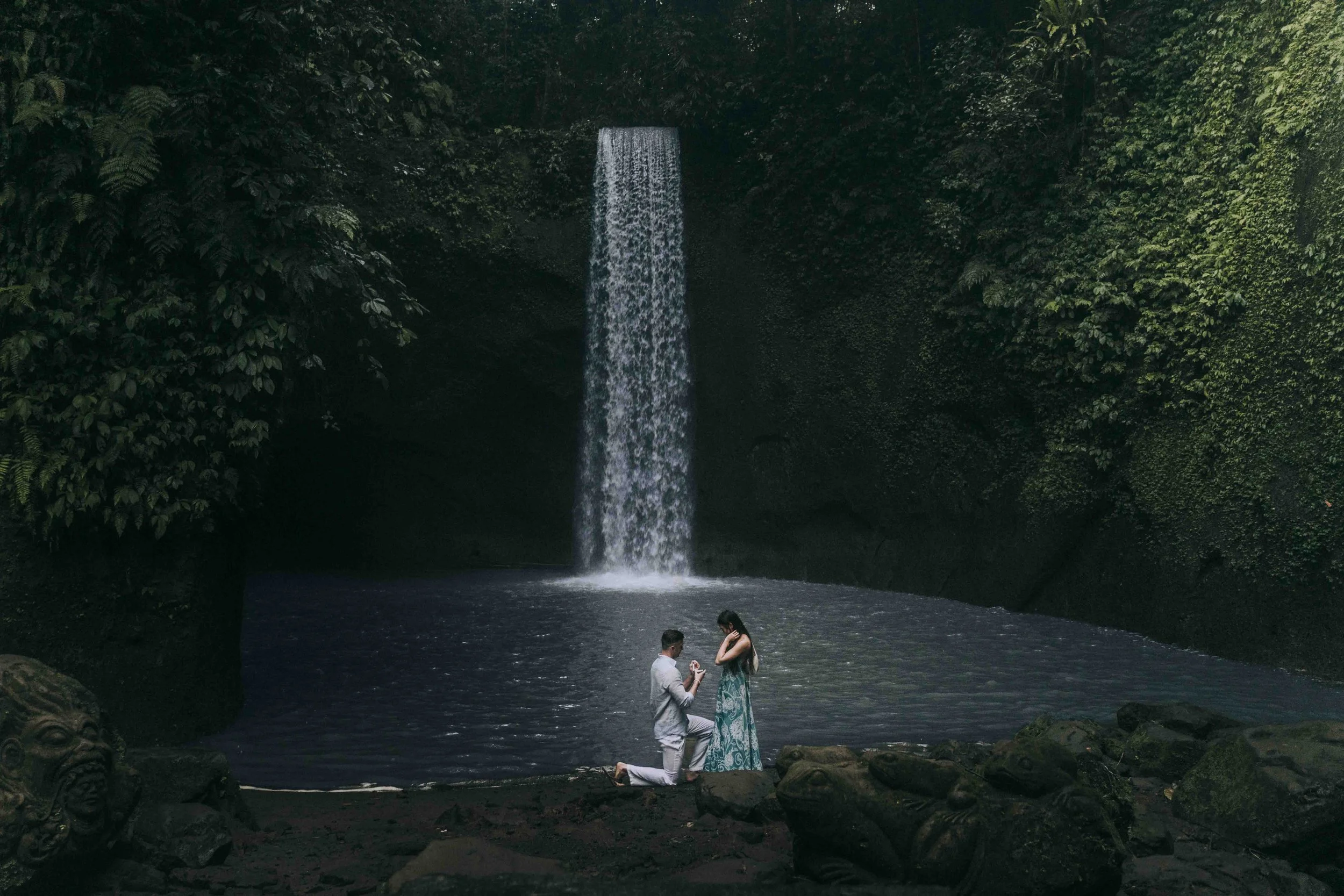 A man kneels beside a still pool while a woman stands facing him, both centered beneath a waterfall, observed by a Fragmented Memories photographer.