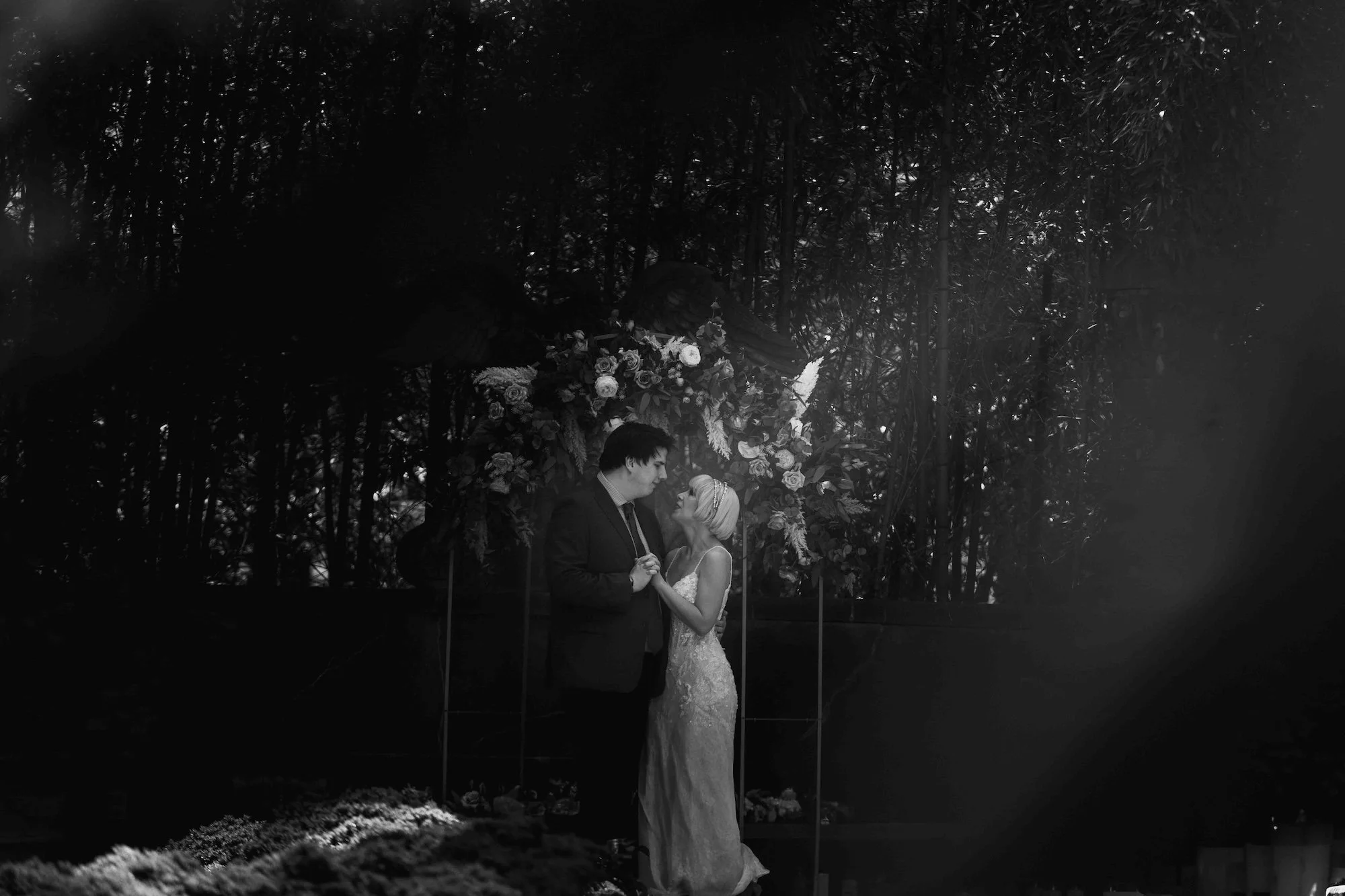Cinematic kiss under dappled light, the couple framed by trees in a quiet setting.