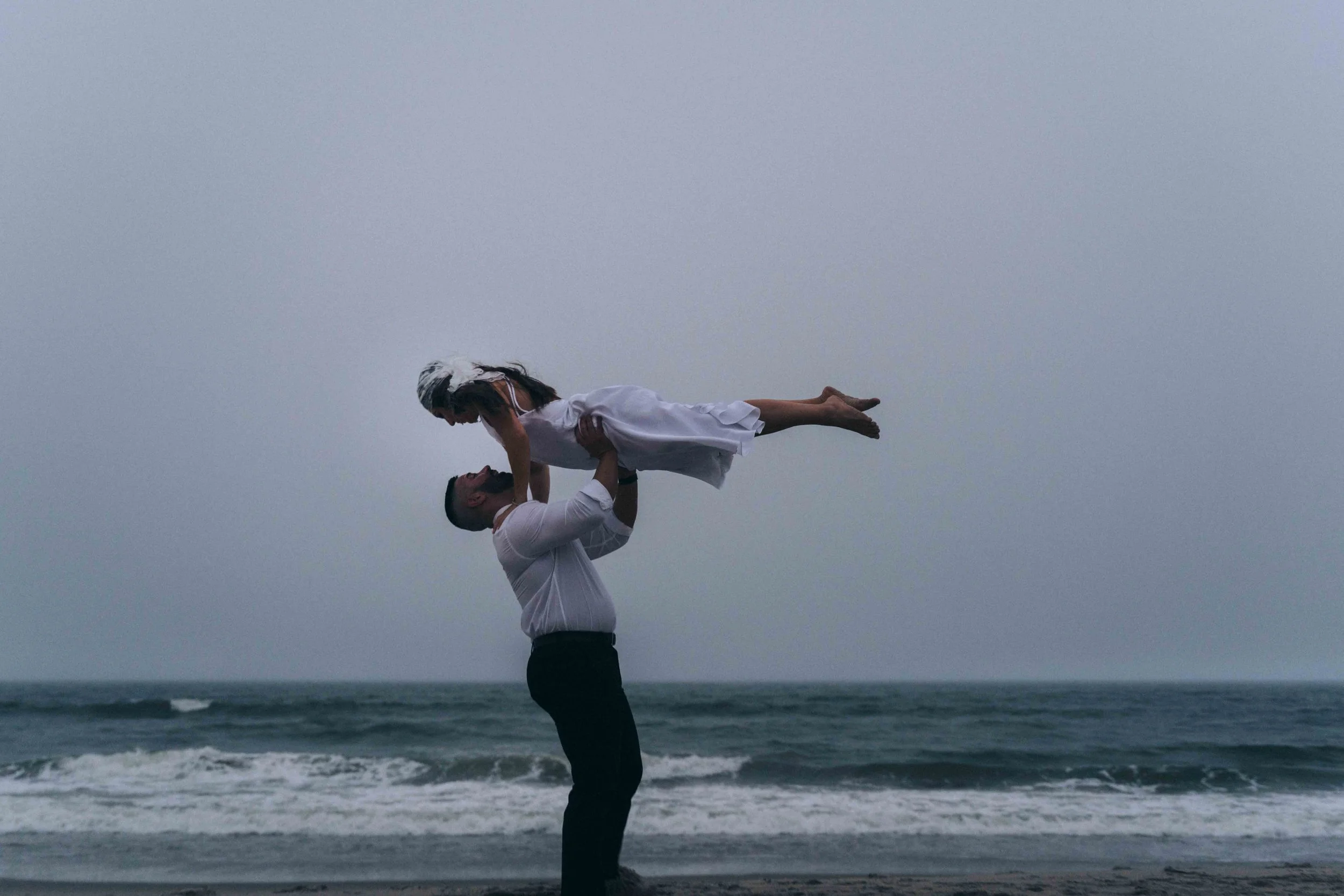 A man lifts a woman into the air at the shoreline as waves roll in behind them, captured as a Fragmented Memories prenup photographer frames the moment.
